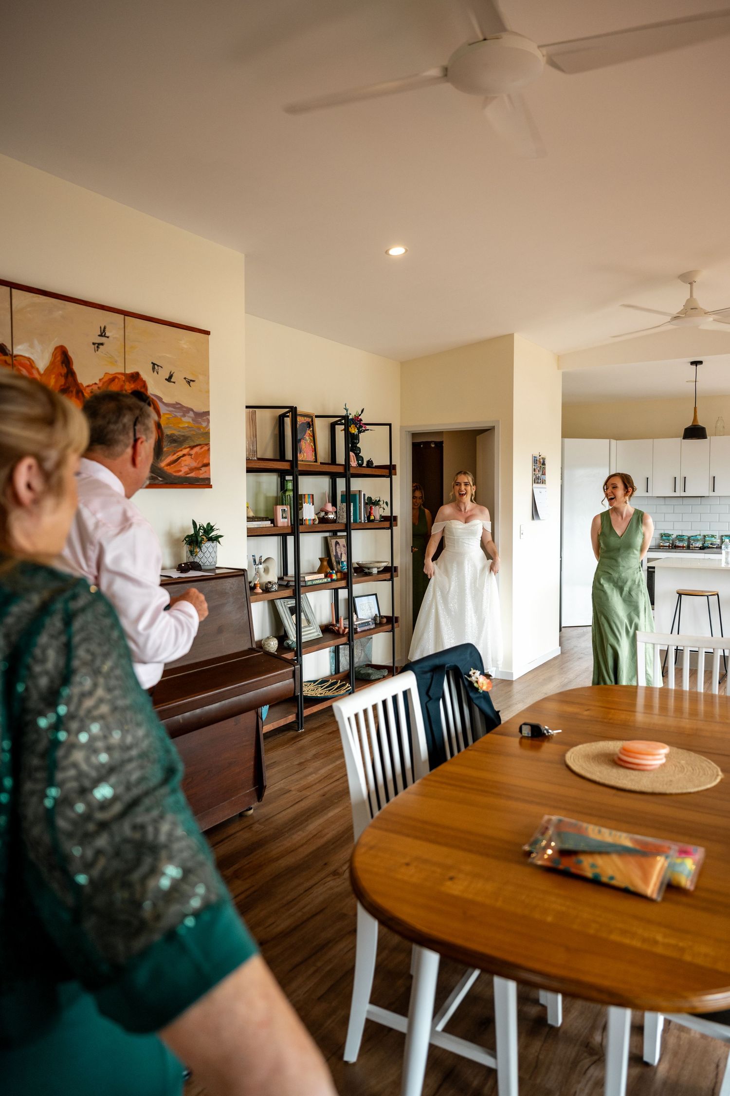 Bride in white wedding gown making first appearance to family in bright open-plan home interior.