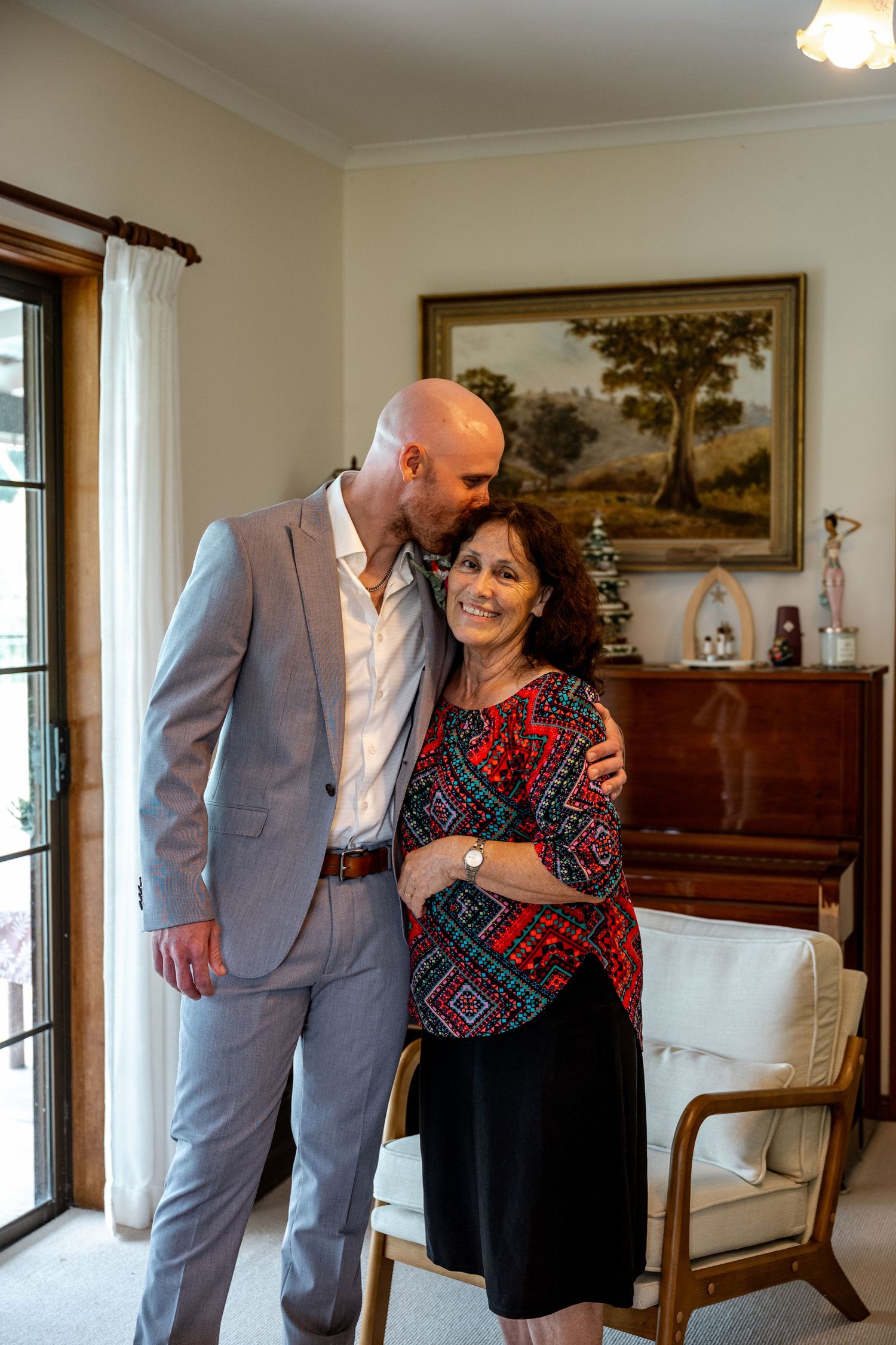 Bald groom in grey suit kissing mother on cheek while embracing in elegant living room.