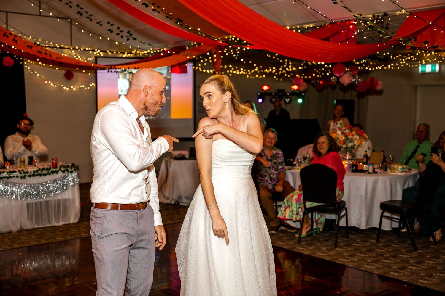 Groom in white shirt dances with smiling bride in strapless gown at a decorated wedding reception.