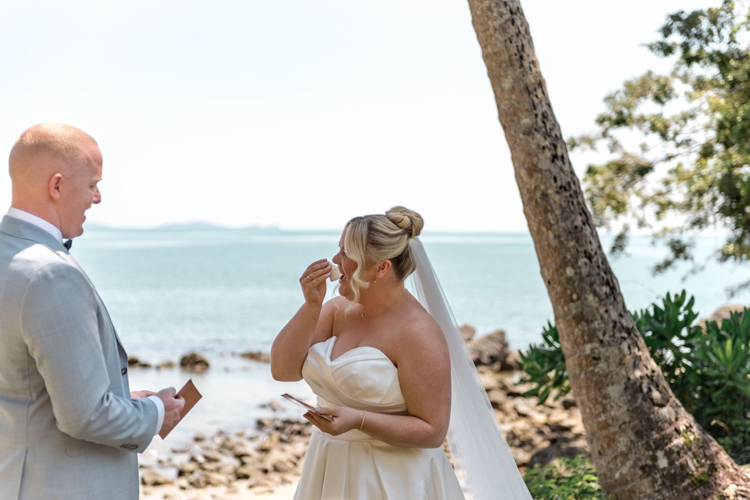 Emotional bride wipes tears reading vows at beachside wedding ceremony under palm trees.