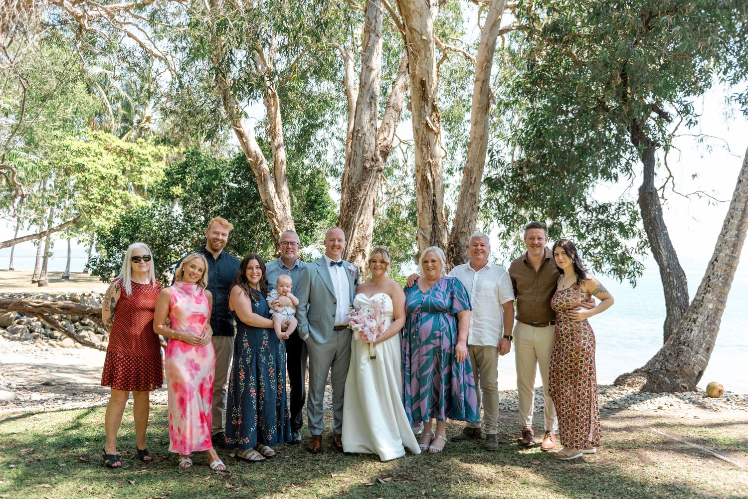 Wedding party group photo of ten guests with bride and groom holding baby under eucalyptus trees by water.