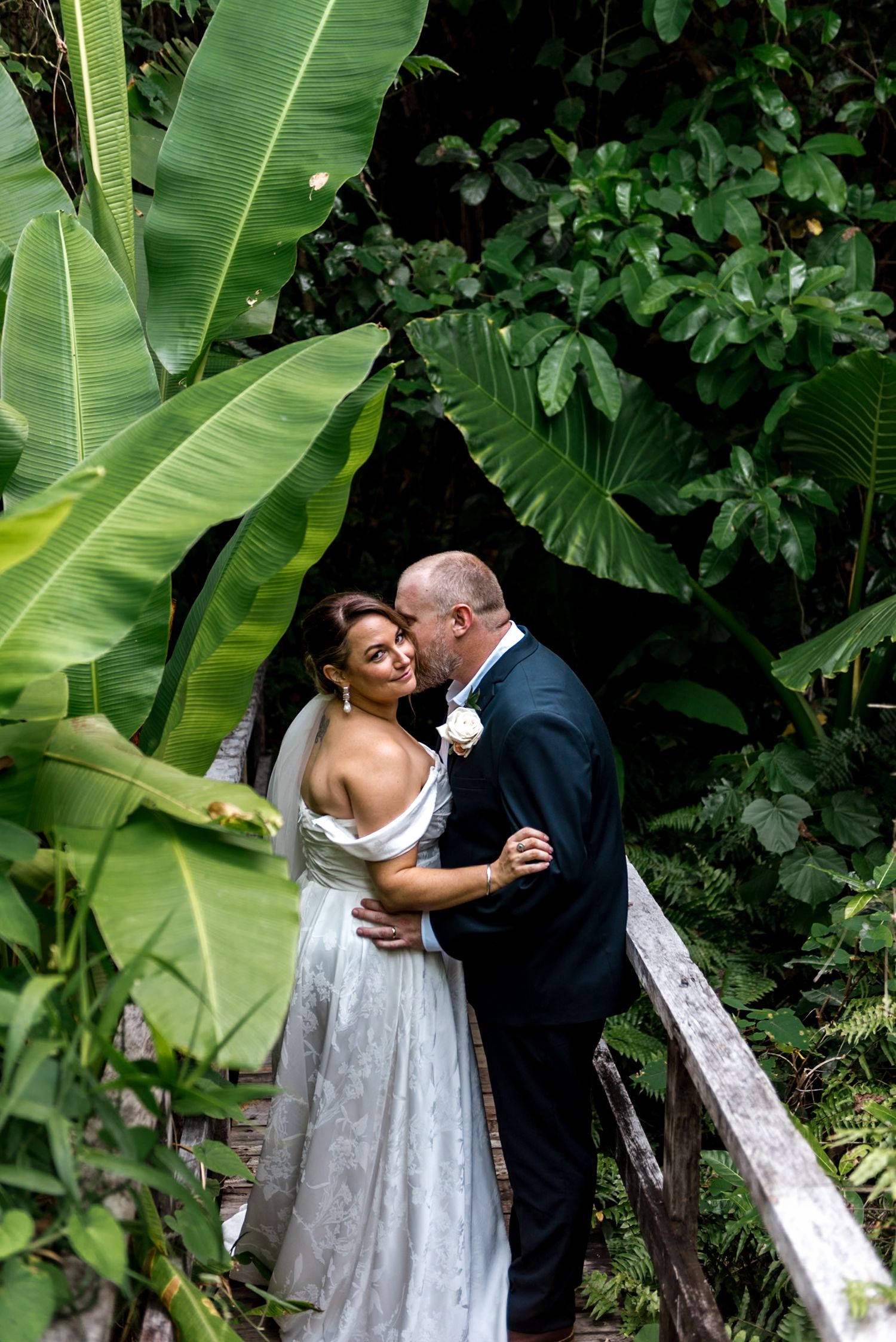 Groom kisses bride on cheek as they stand on wooden bridge surrounded by tropical foliage.