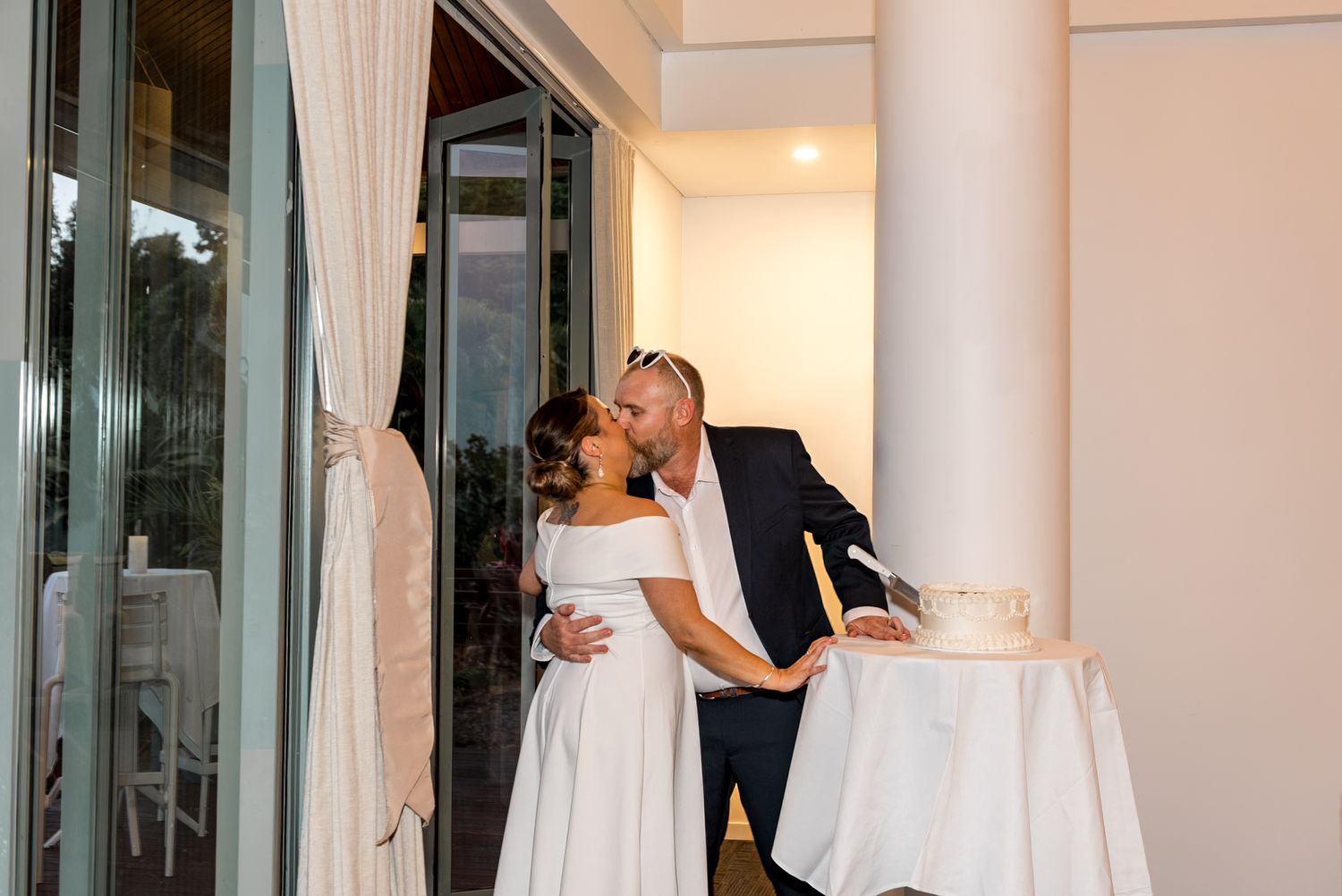 Bride and groom sharing a romantic kiss beside their white wedding cake at the reception.