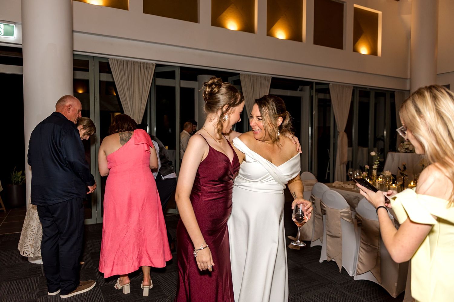 Bride in white dress shares a joyful moment with a bridesmaid on dance floor.