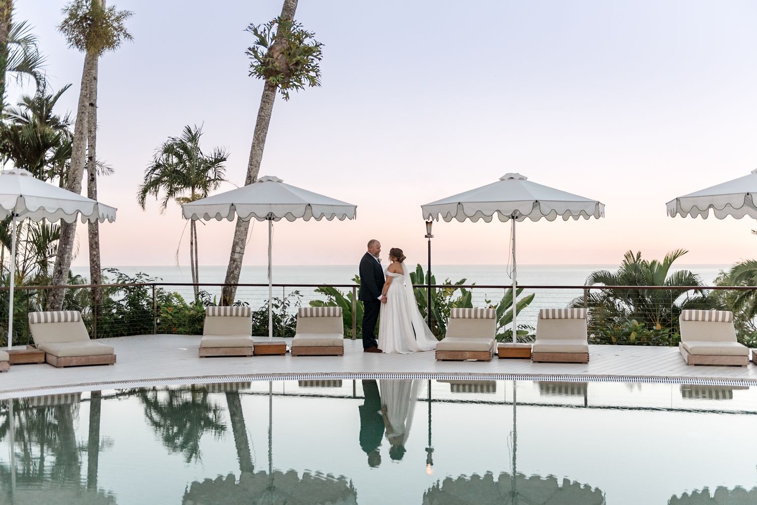 Bride and groom share a romantic moment by a resort pool at sunset with ocean views and white umbrellas.