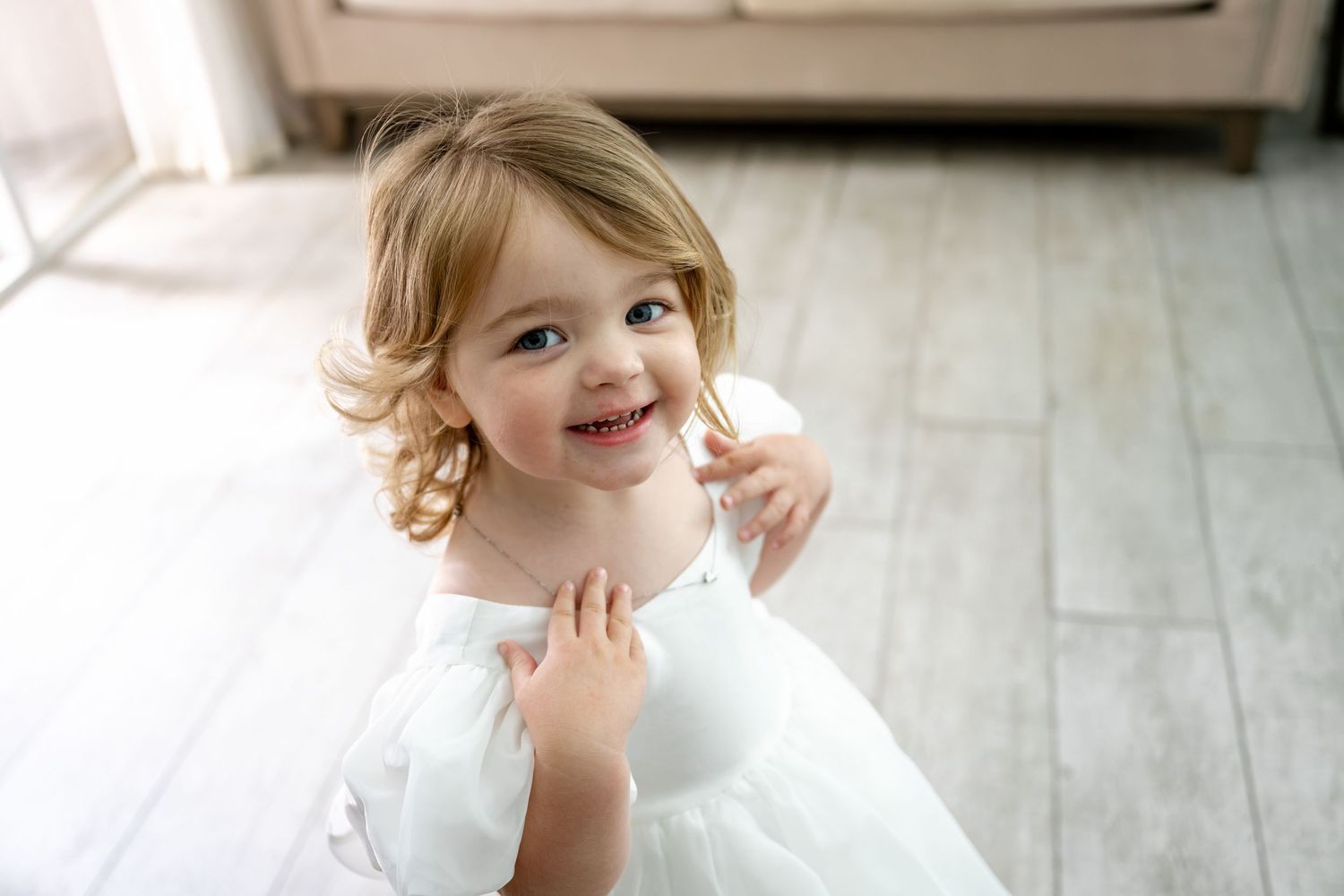 Cute smiling toddler girl in a white flower girl dress looking up at the camera on a wood floor.