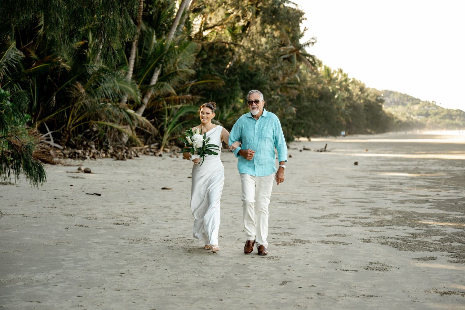 Elegant couple strolling along sandy tropical beach, woman in white outfit holding green bouquet, man in turquoise shirt at Four Mile Beach.