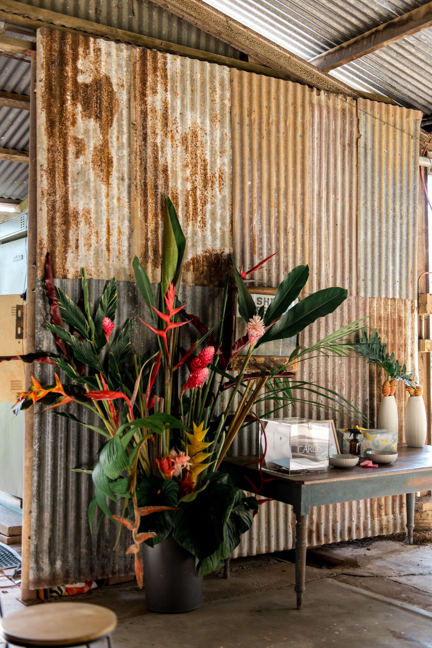 Large tropical floral arrangement with birds of paradise and heliconia against rustic corrugated iron barn wall.