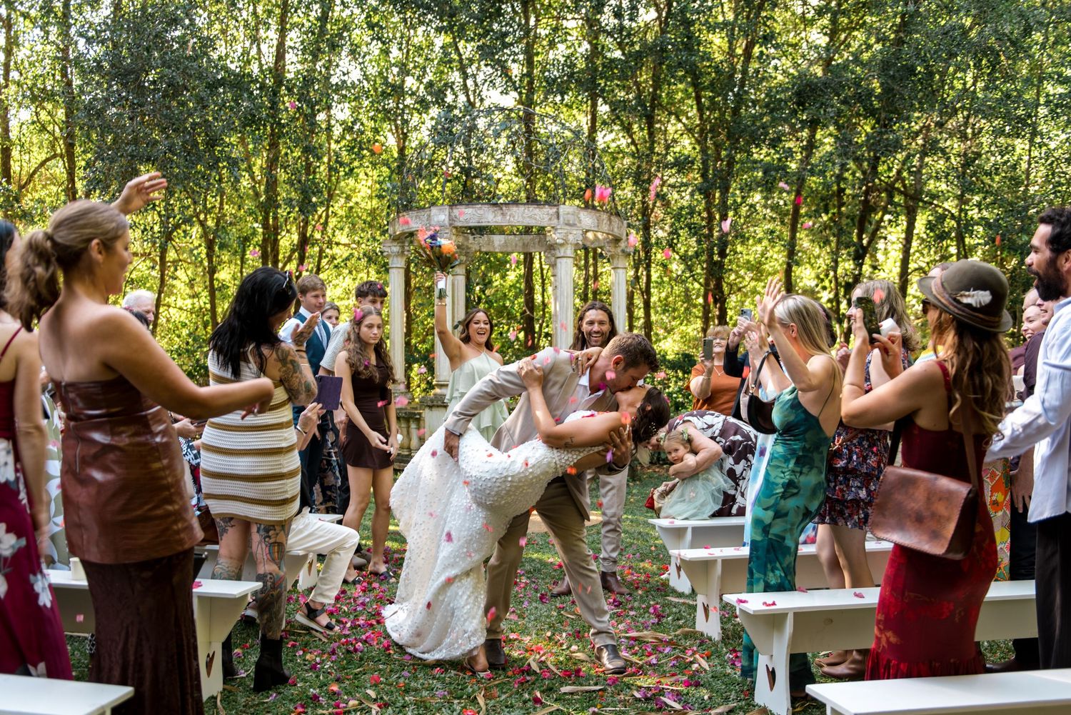 Groom dips bride in celebratory kiss as wedding guests cheer along the aisle after outdoor ceremony.