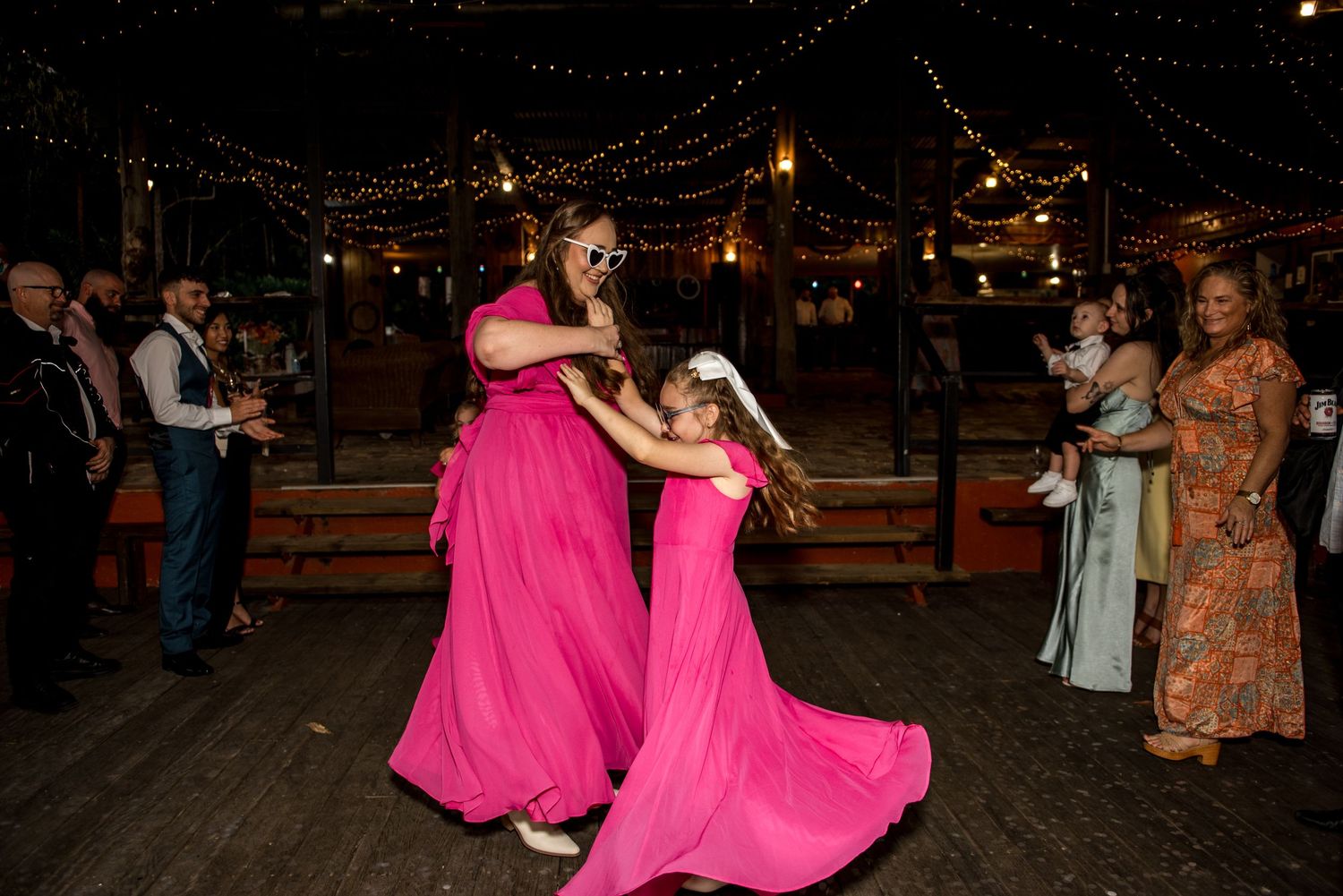 Two guests in bright pink gowns dance joyfully under string lights at an outdoor wedding reception.