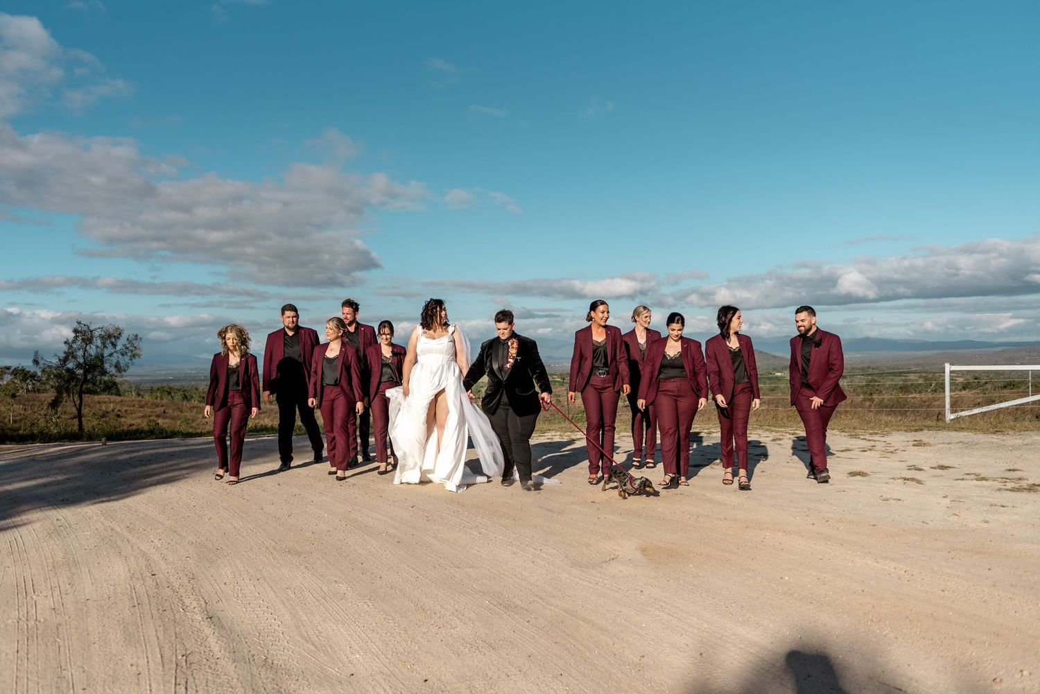 Same-sex wedding party in burgundy suits walking along a dusty outback road with panoramic Australian countryside views.
