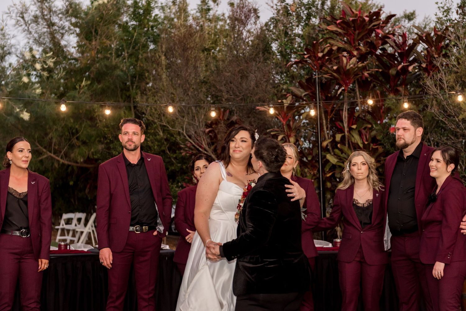 Bride shares emotional hug with guest while wedding party in burgundy suits stands nearby.
