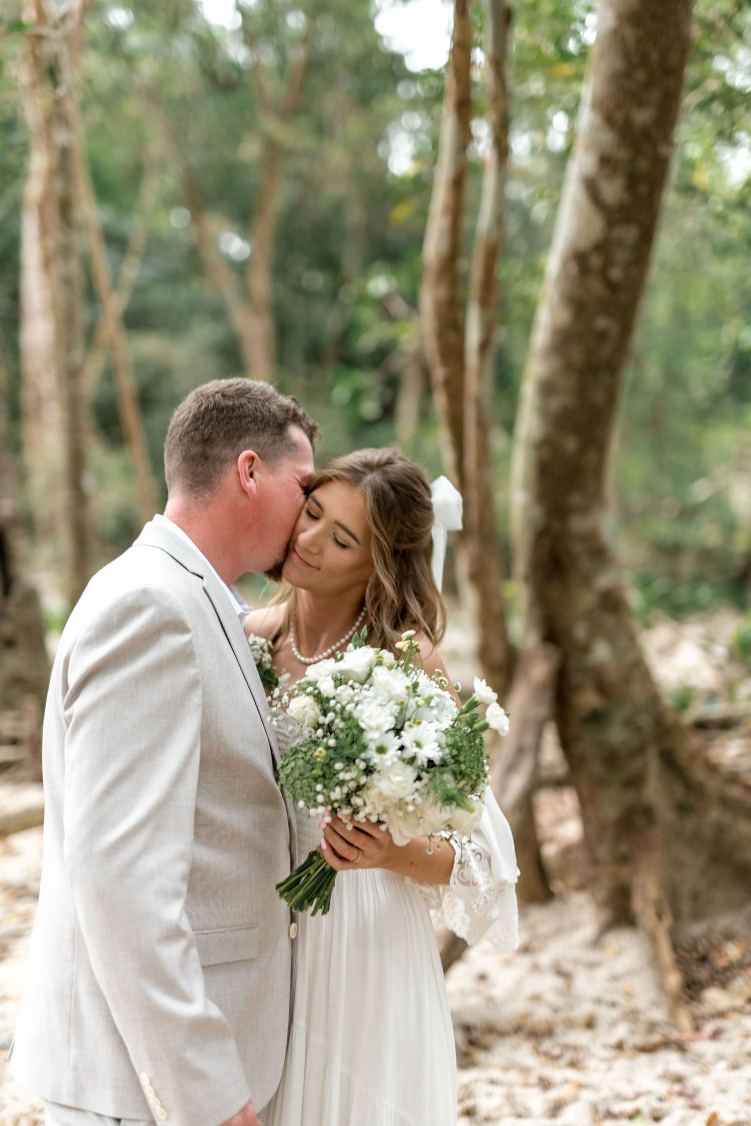 Groom kisses bride on the cheek as she holds a white floral bouquet in a woodland setting.
