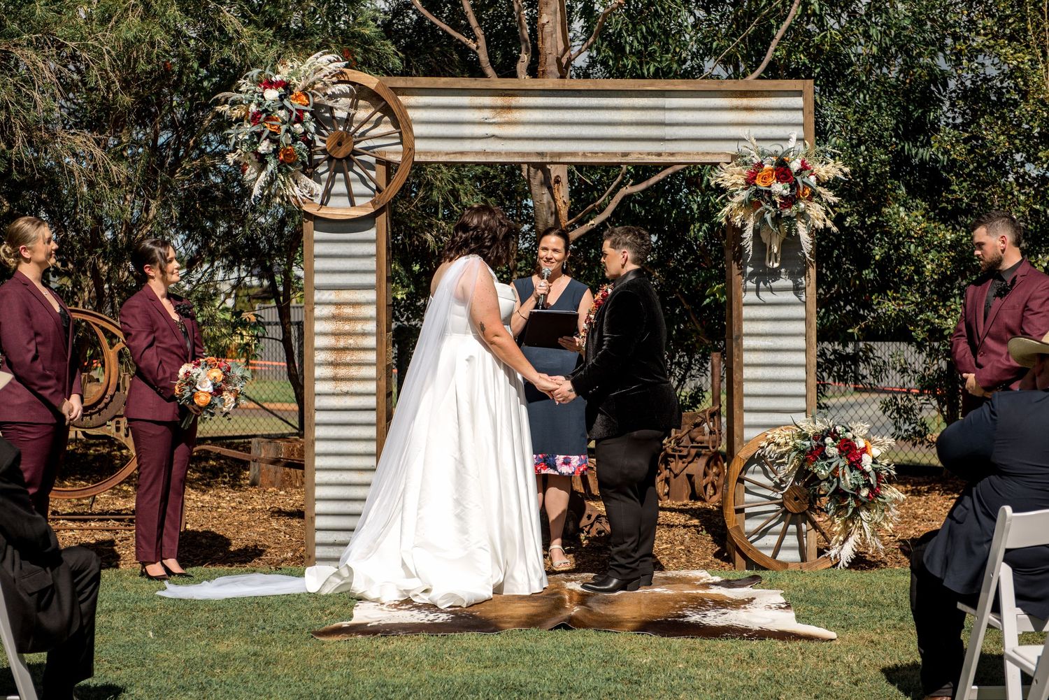 Couple exchanges vows beneath rustic wagon wheel arch adorned with vibrant flowers at outdoor wedding ceremony.