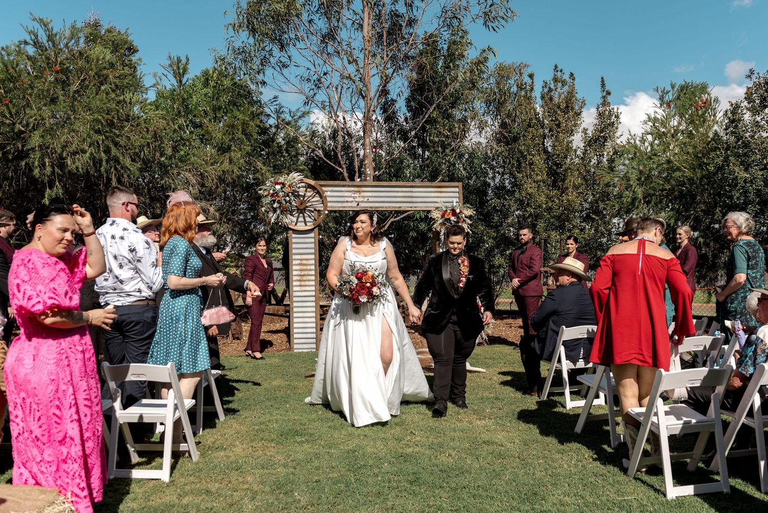 Bride and groom exit wedding ceremony down aisle as guests celebrate at outdoor garden venue.