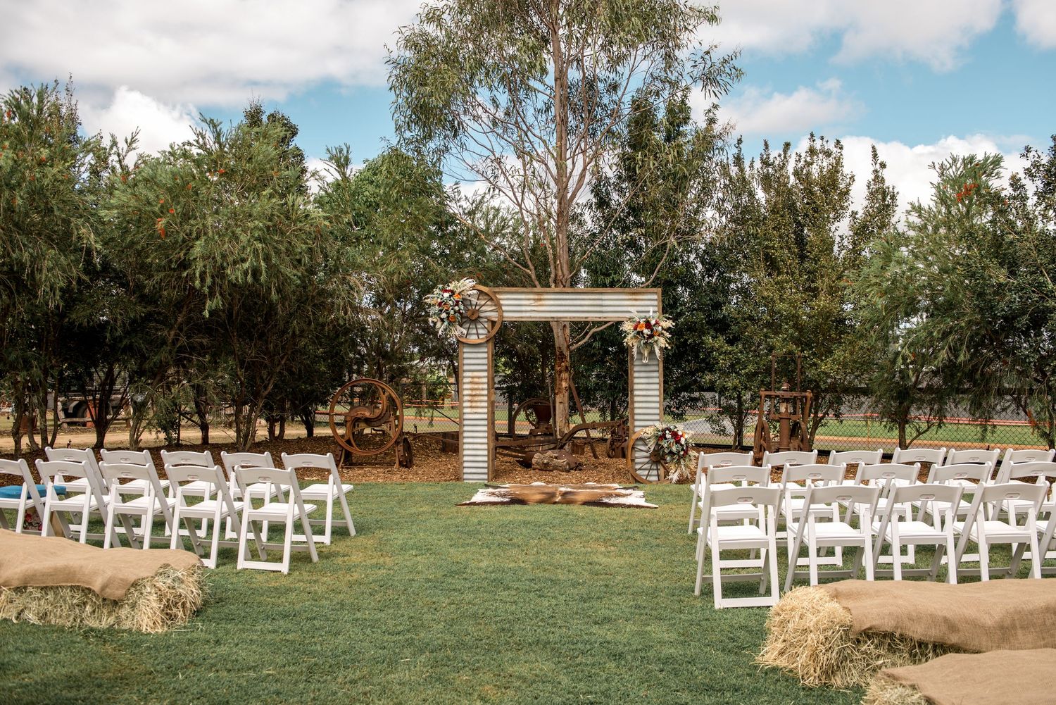Rustic outdoor wedding ceremony setup with white chairs, hay bales, and a wooden arch decorated with flowers on green lawn.