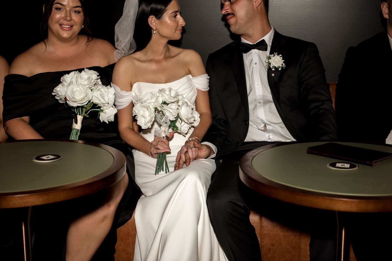 Bride in off-shoulder white gown sits between bridesmaids in black dresses holding white roses.