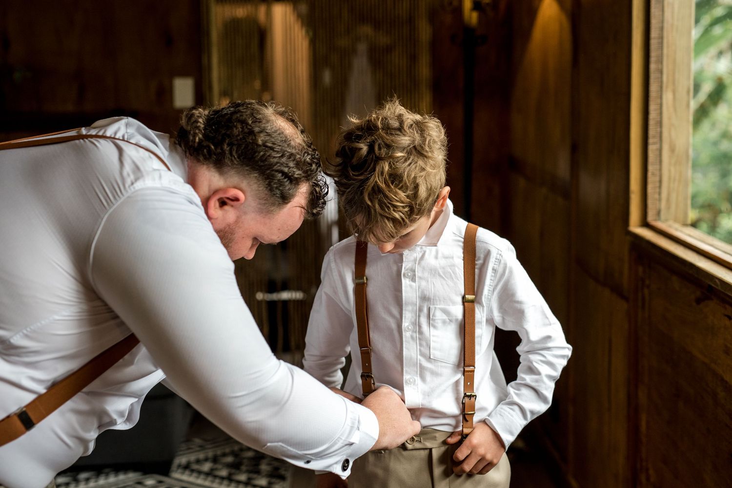Groom in white shirt and suspenders helping young boy fasten his brown suspenders indoors.
