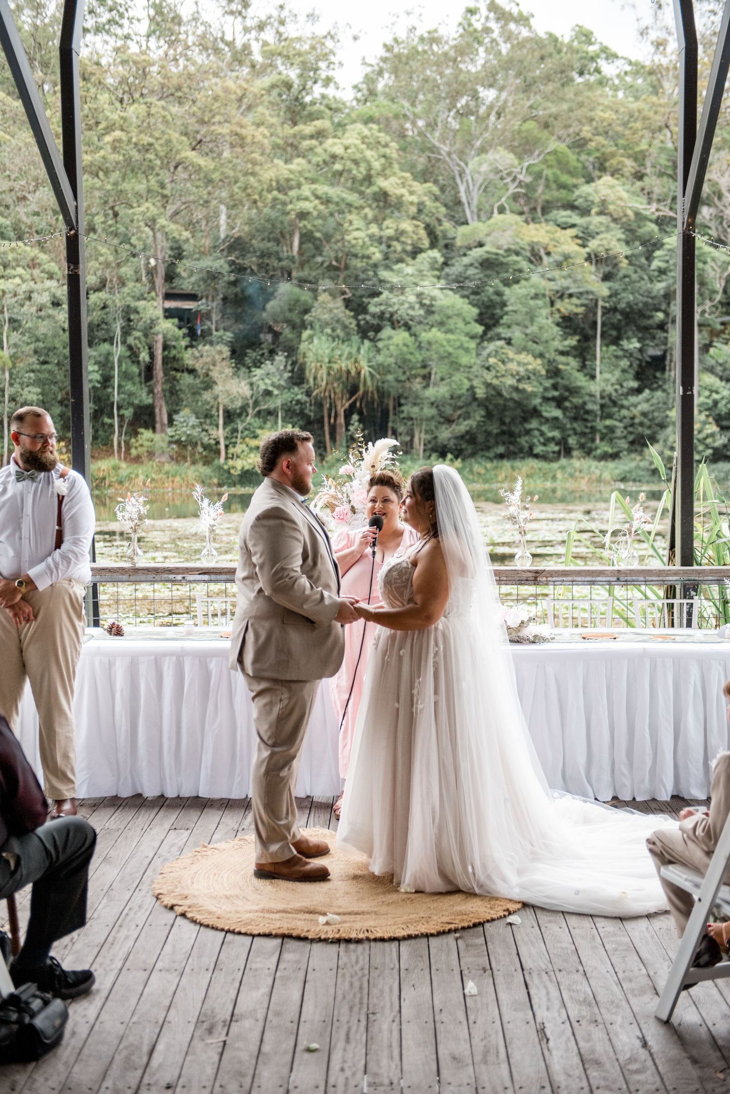 Bride and groom exchange vows at outdoor wedding ceremony with officiant, lush green forest backdrop visible.