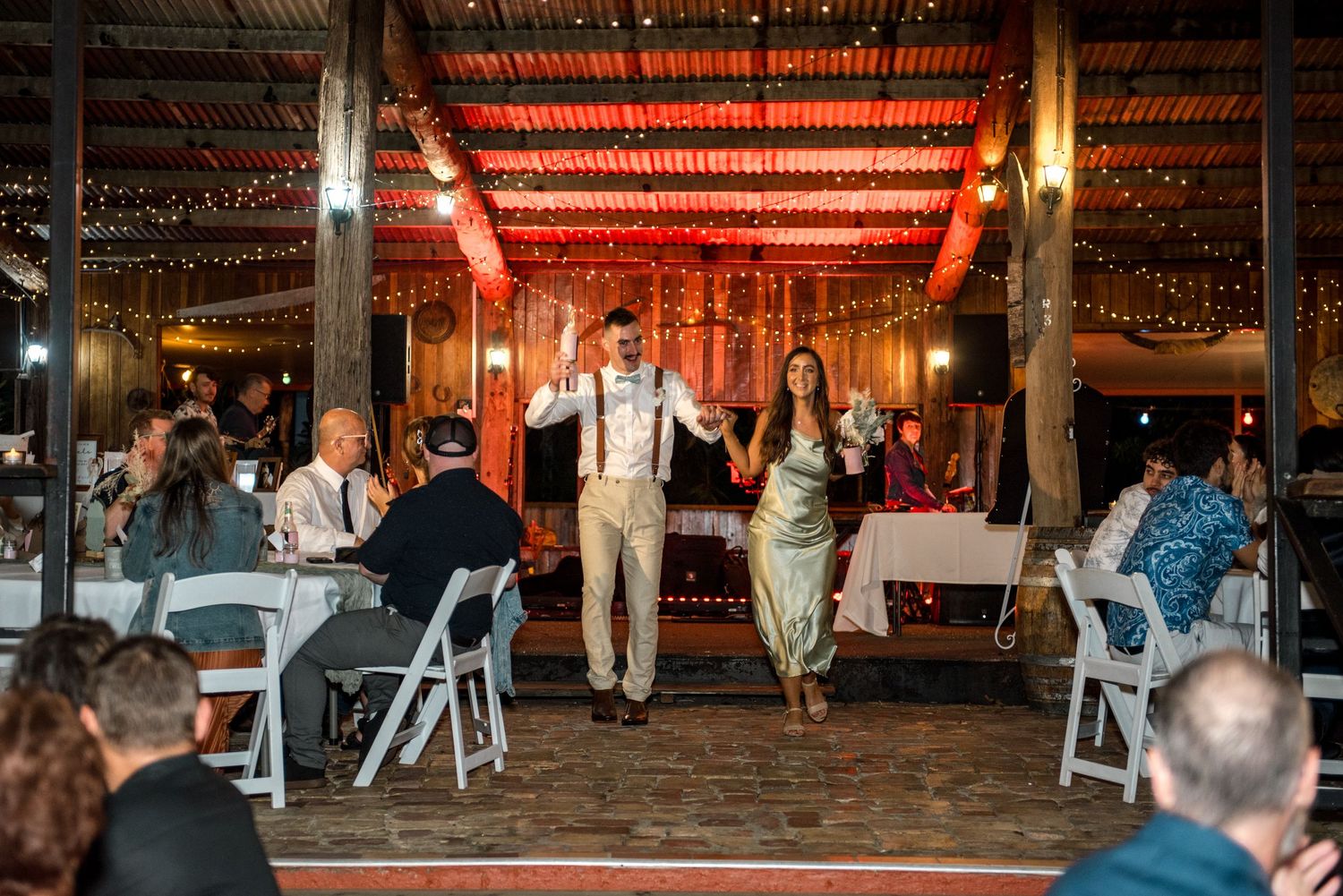 Bride and groom walk hand in hand through decorated barn wedding reception with fairy lights overhead.