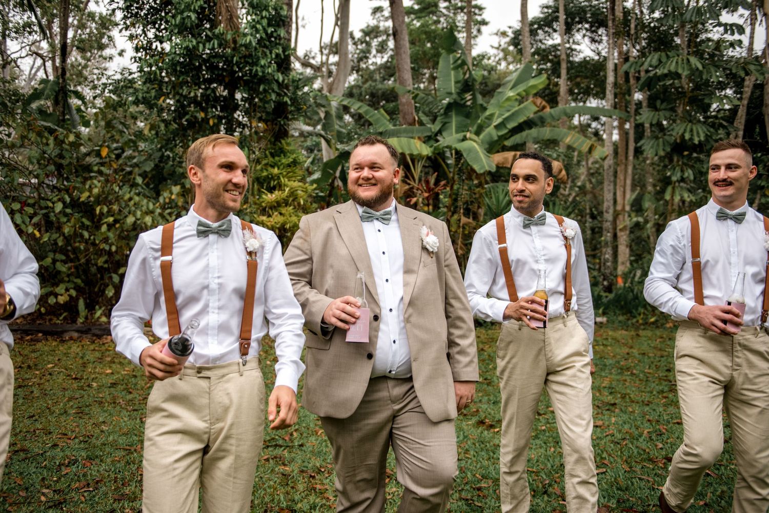 Groom and groomsmen walk casually holding drinks in a lush green garden during wedding celebrations.
