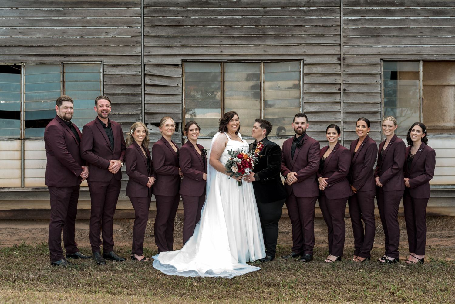 Bride and groom pose with full wedding party in matching burgundy suits outside rustic wooden building.