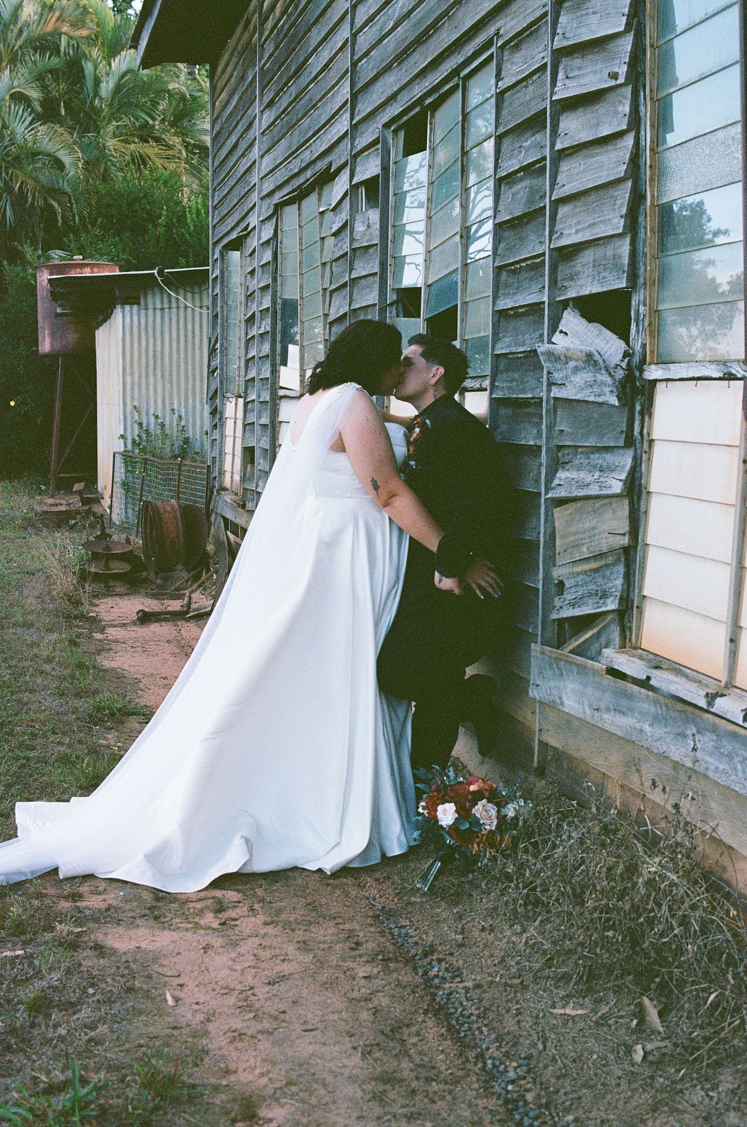 Bride and groom share a romantic kiss against a rustic weathered wooden building on their wedding day.