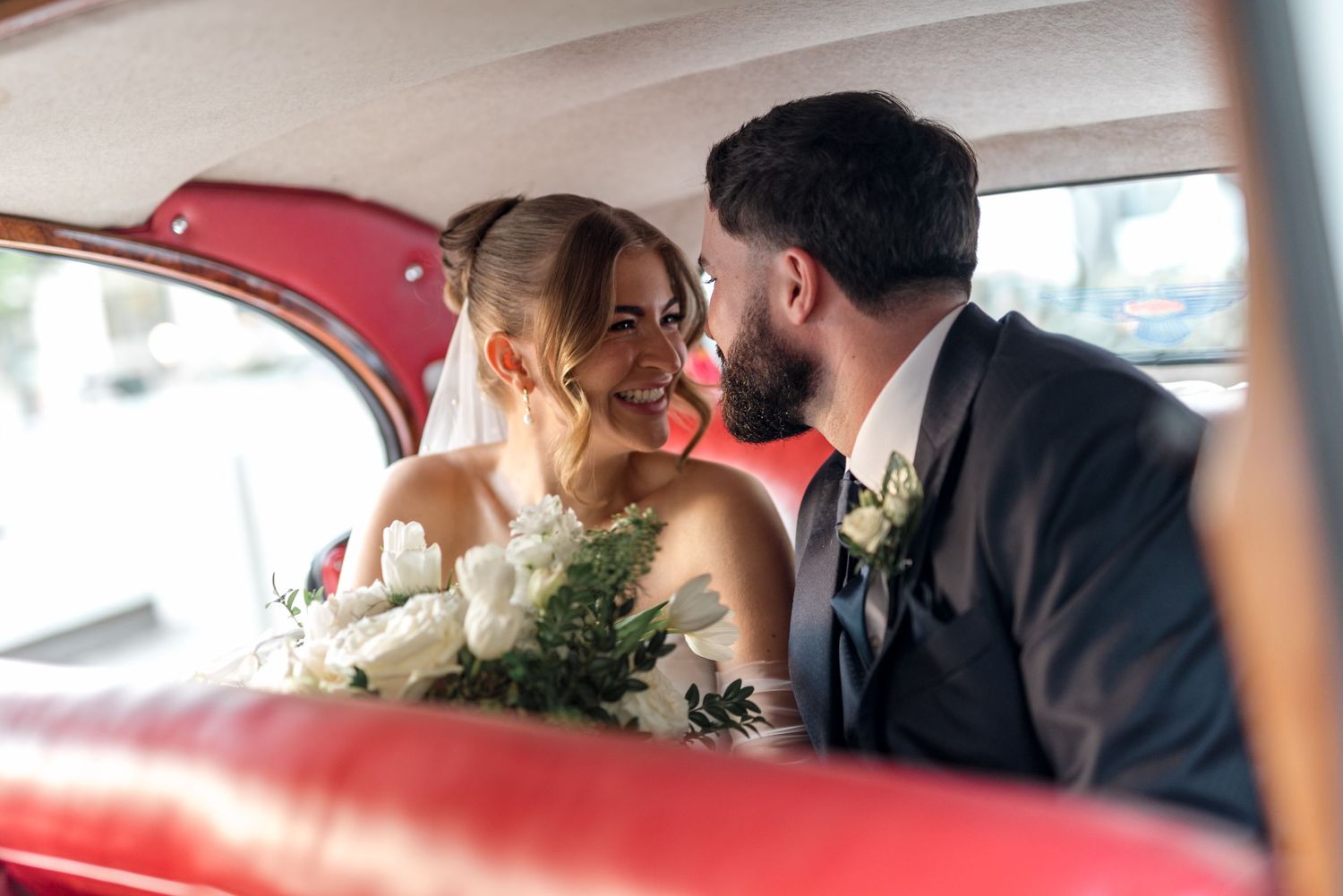 Smiling bride and groom share an intimate moment inside a classic car with red leather seats.