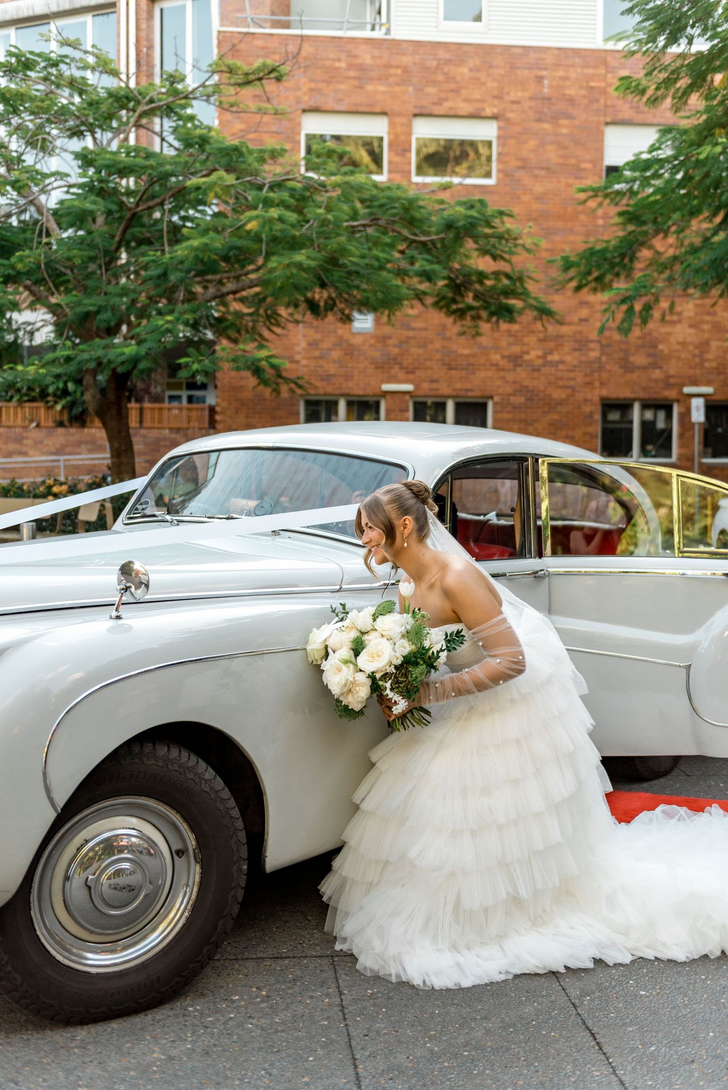 Bride in layered white tulle gown leans gracefully against a vintage white car.