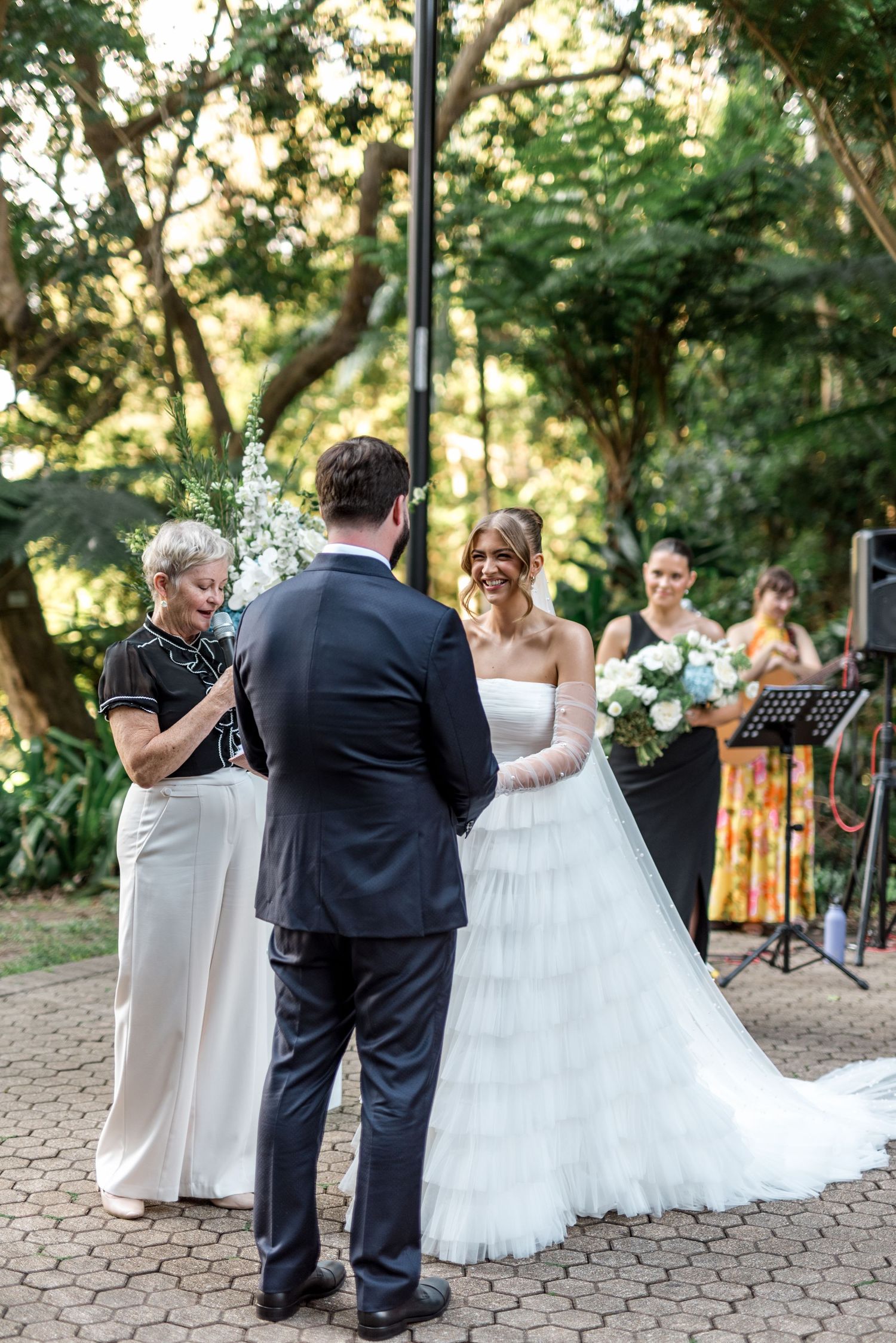Bride in strapless ruffled gown laughs joyfully at groom during outdoor garden wedding ceremony.