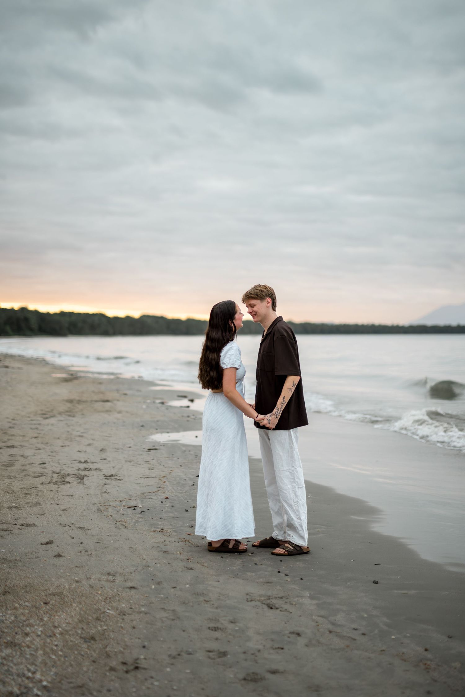 Couple holds hands facing each other on serene beach at cloudy sunset.
