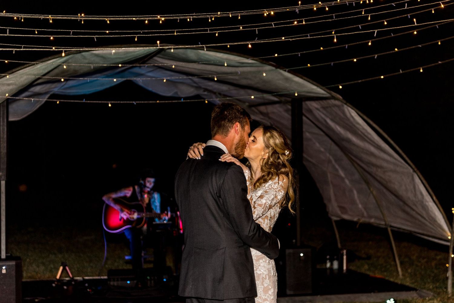 Couple shares their first dance under string lights as a guitarist performs at their outdoor evening wedding.