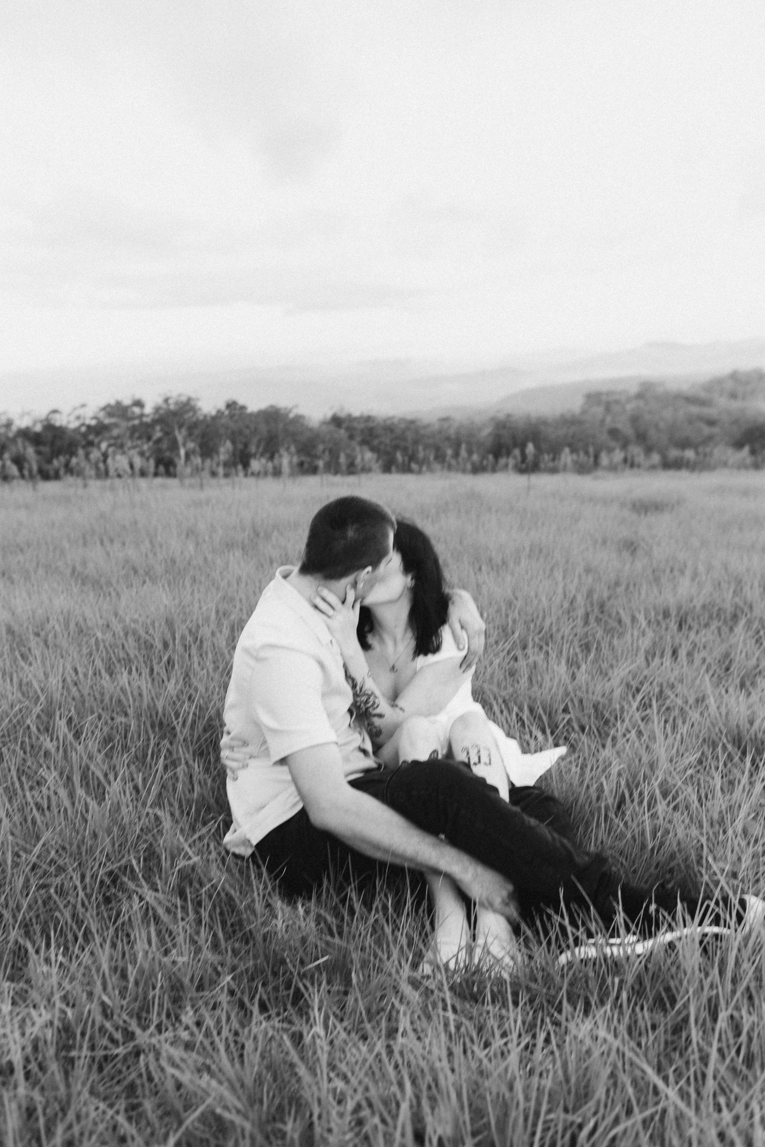 Black and white wide shot of couple kissing in open grassy field with mountain scenery.