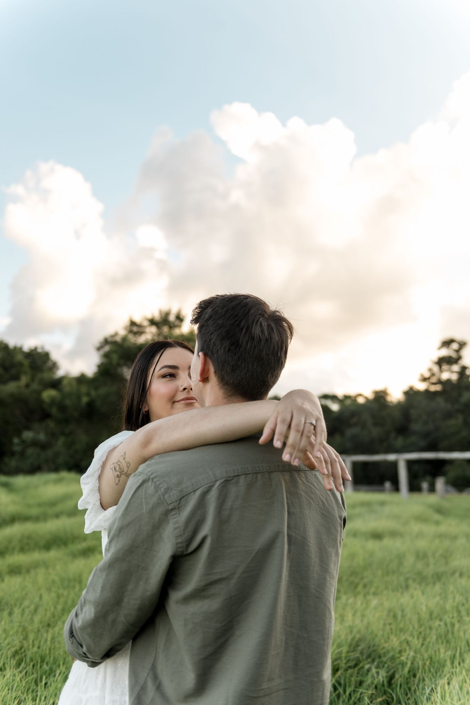 Smiling woman in white dress hugging man in olive shirt in green field with dramatic cloudy sky.