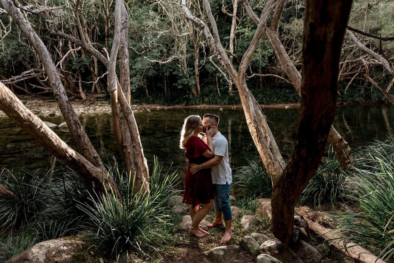 Couple kissing romantically in a forest setting beside a calm creek surrounded by native trees.