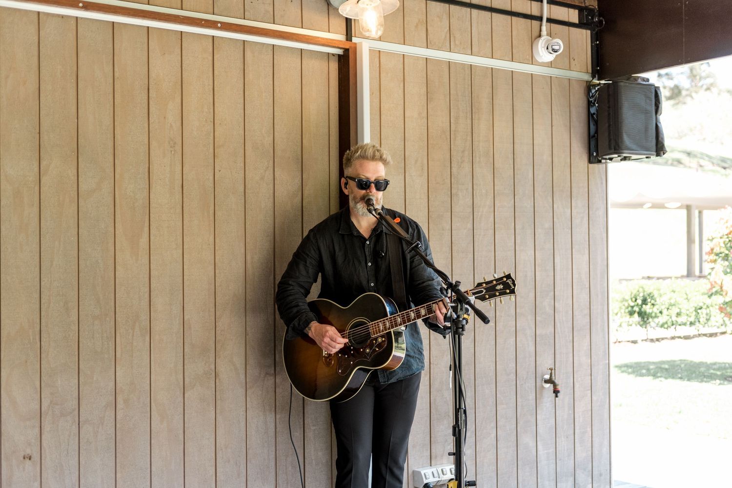 Male guitarist performing acoustic music at an outdoor wedding ceremony against a wooden shiplap wall backdrop.