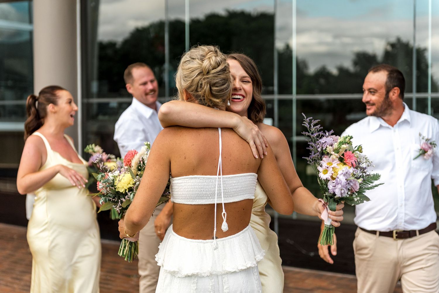 Bride hugs a bridesmaid in a yellow dress while guests celebrate at an outdoor wedding reception.