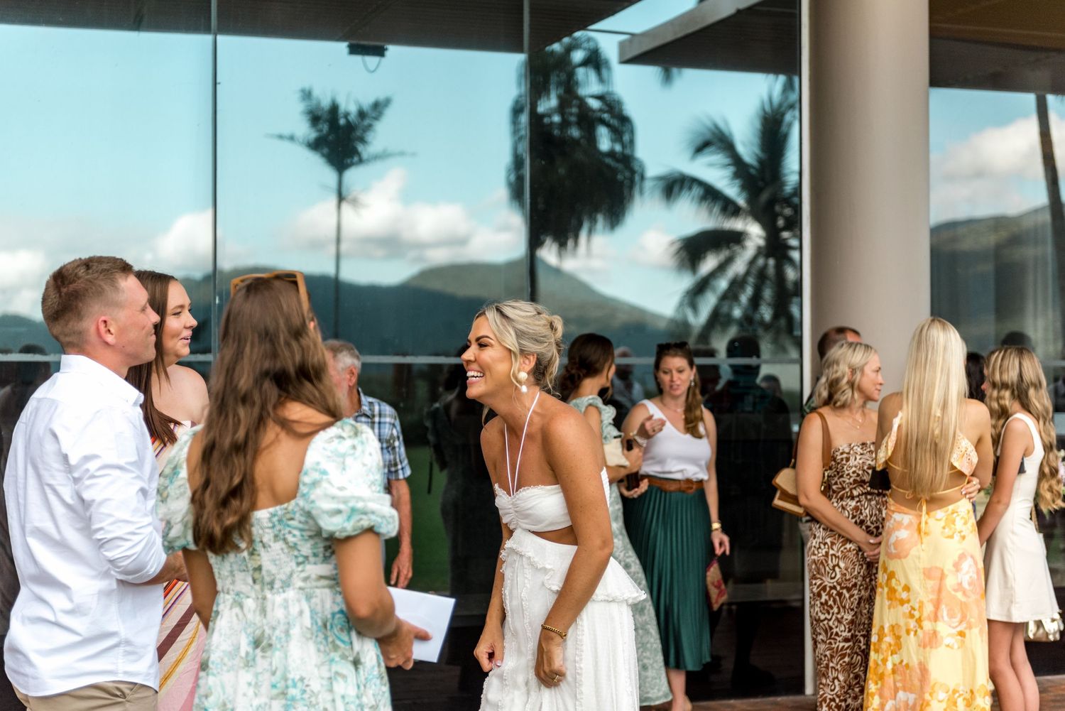Bride in white dress laughs with guests outside a modern glass venue with mountain views.