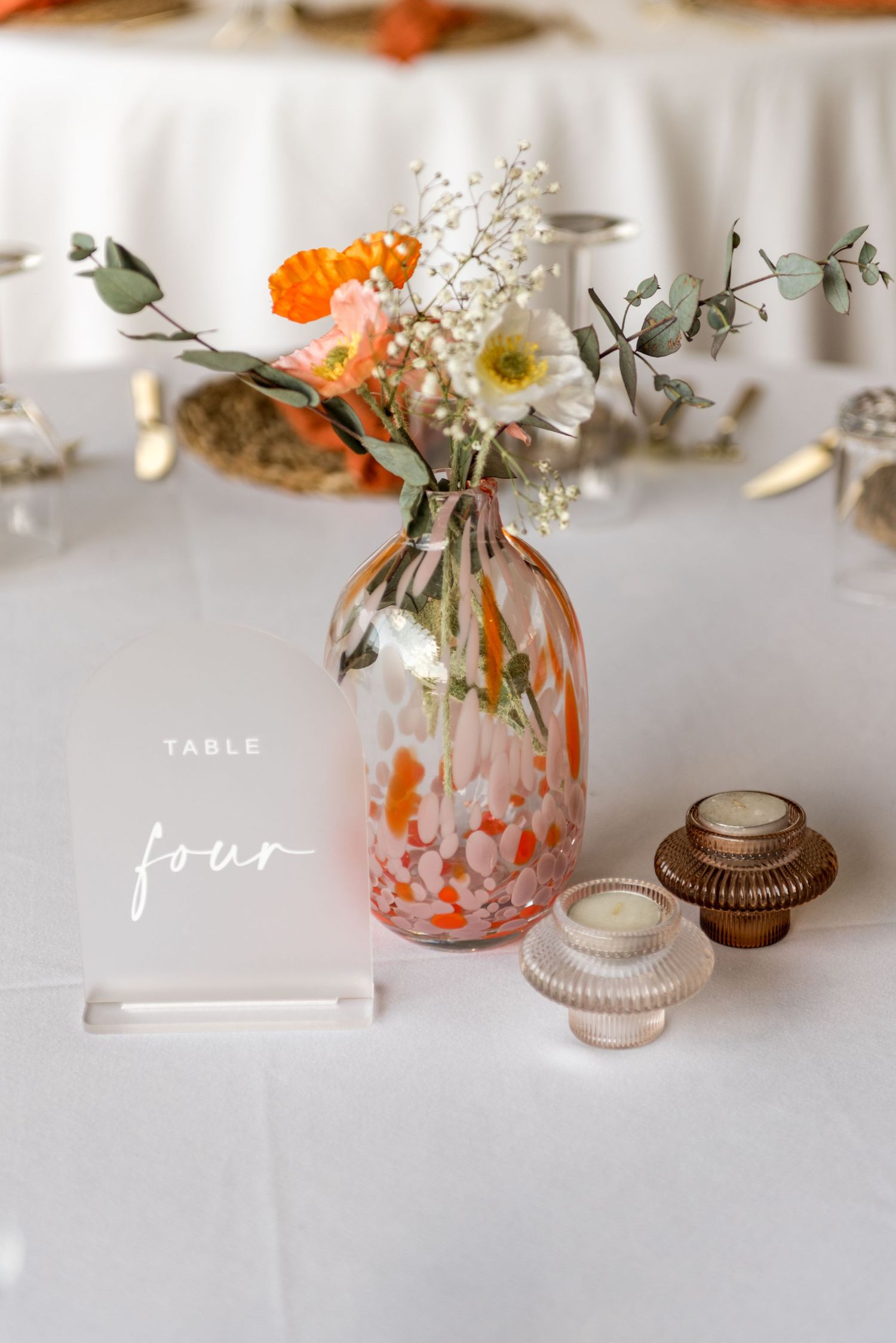 Elegant wedding table four centerpiece with orange speckled glass vase holding wildflowers and baby's breath on white linen.