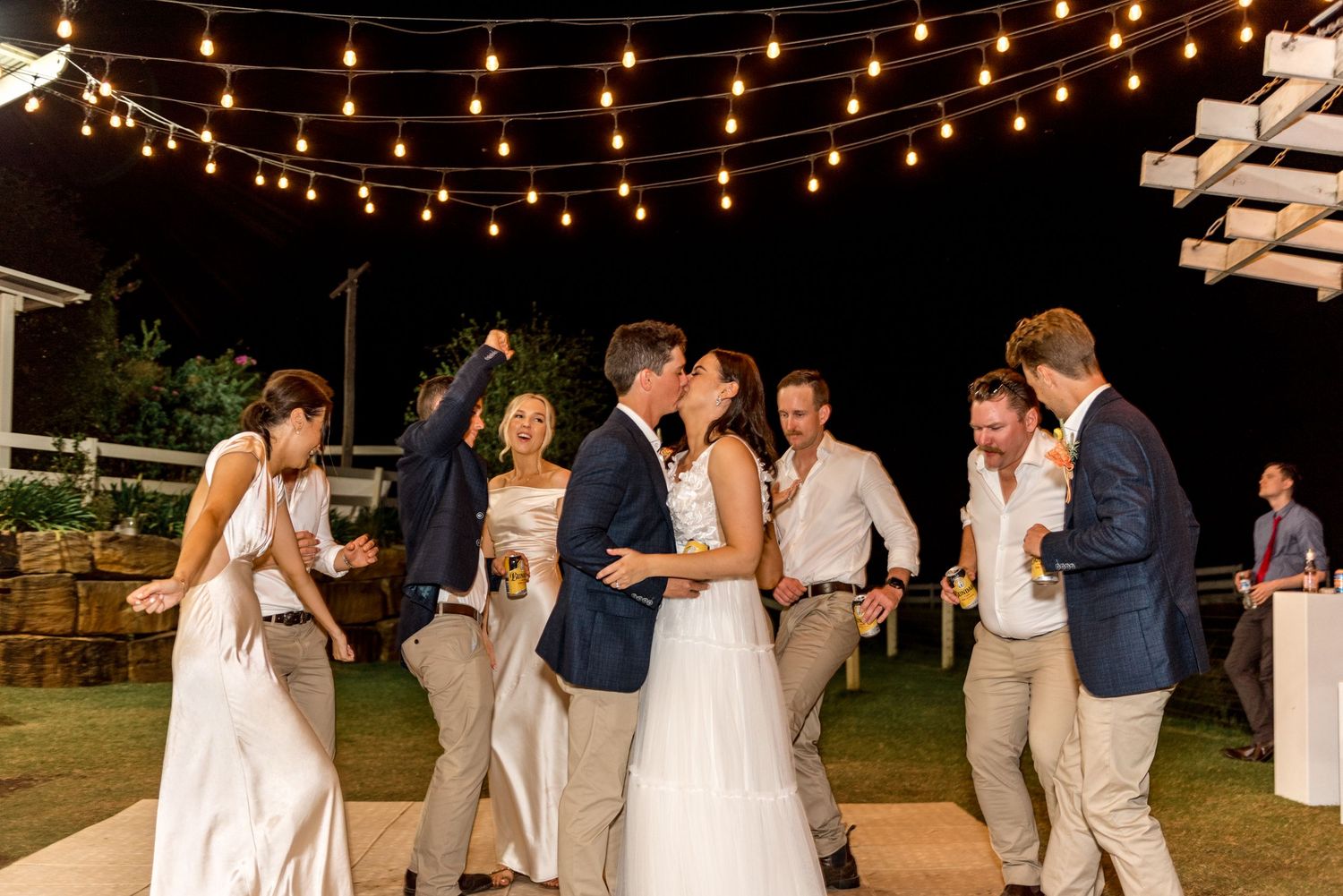 Bride and groom sharing a kiss surrounded by wedding guests dancing under string lights at night.