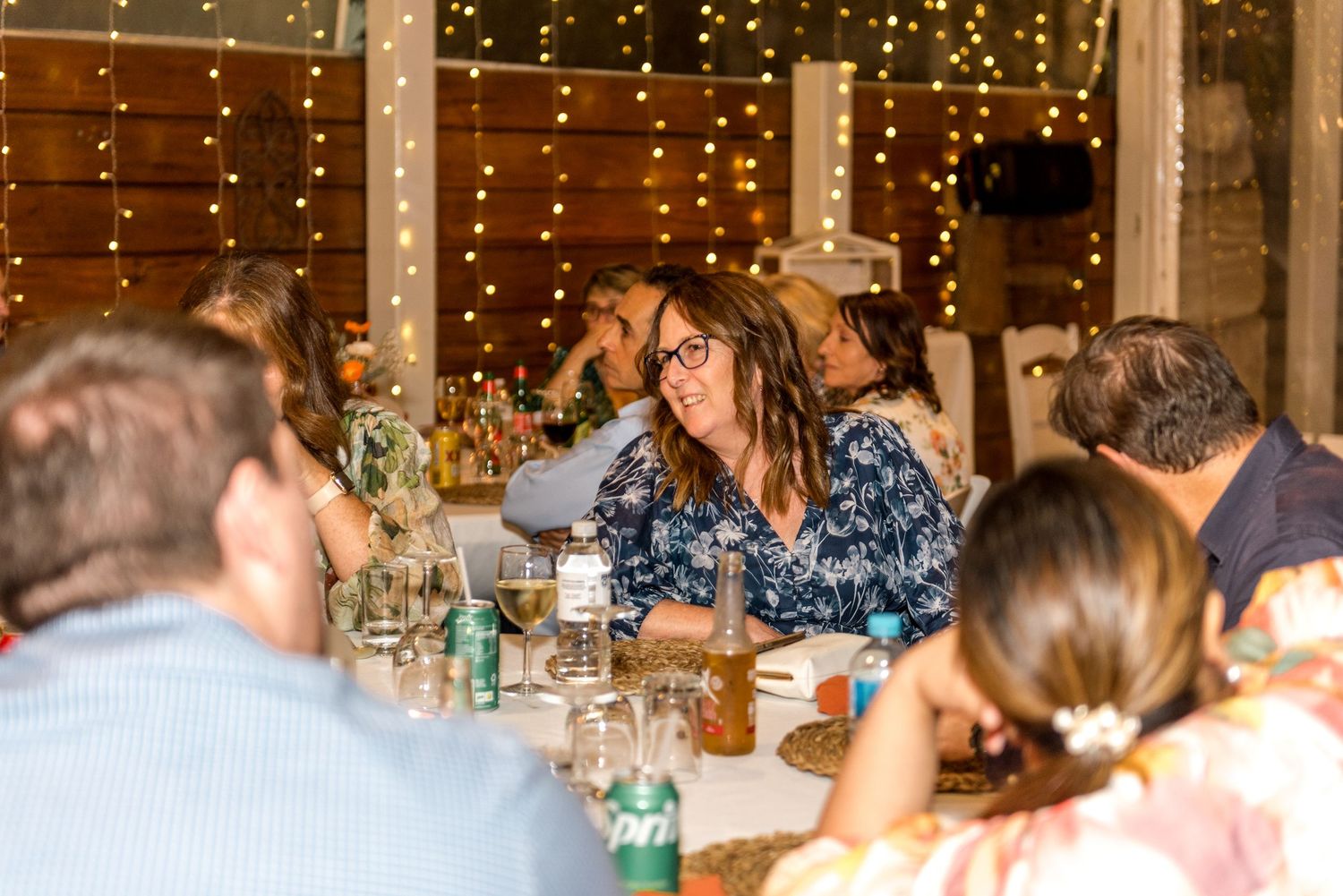 Wedding guests laugh and socialize at long reception tables decorated with fairy lights in a rustic barn.