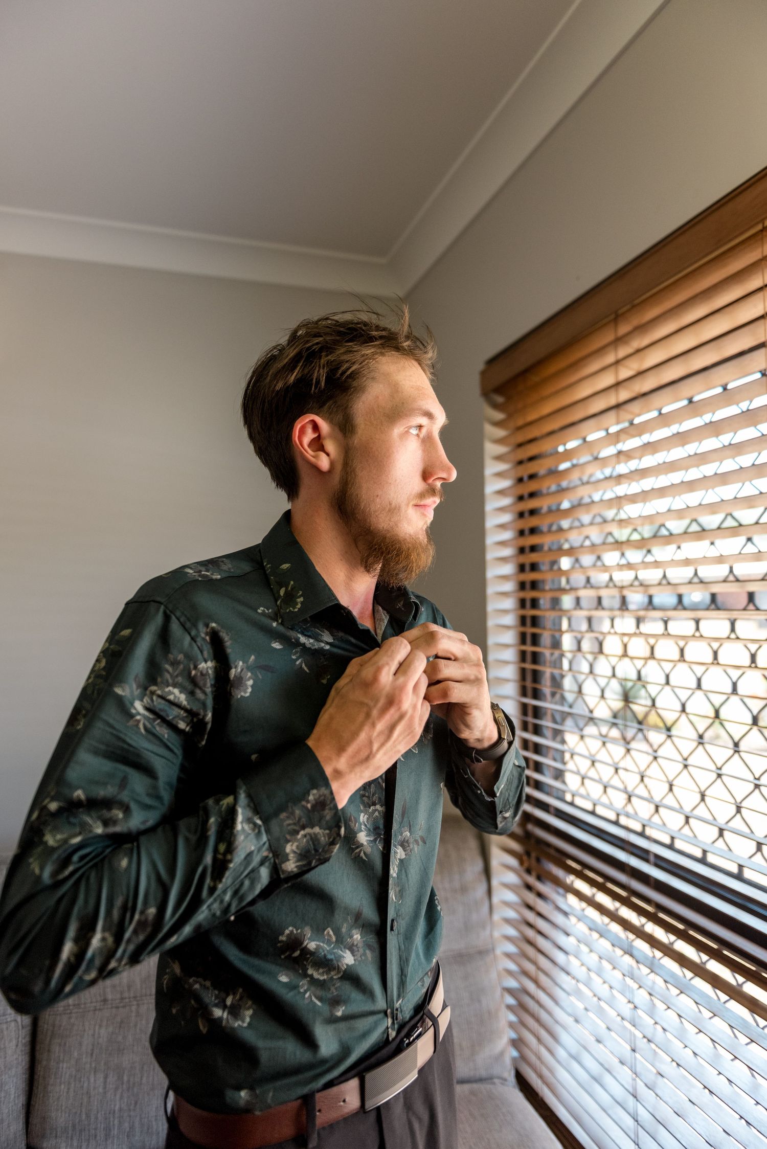 Groom buttoning his dark floral shirt while gazing out a window with wooden blinds on wedding day.