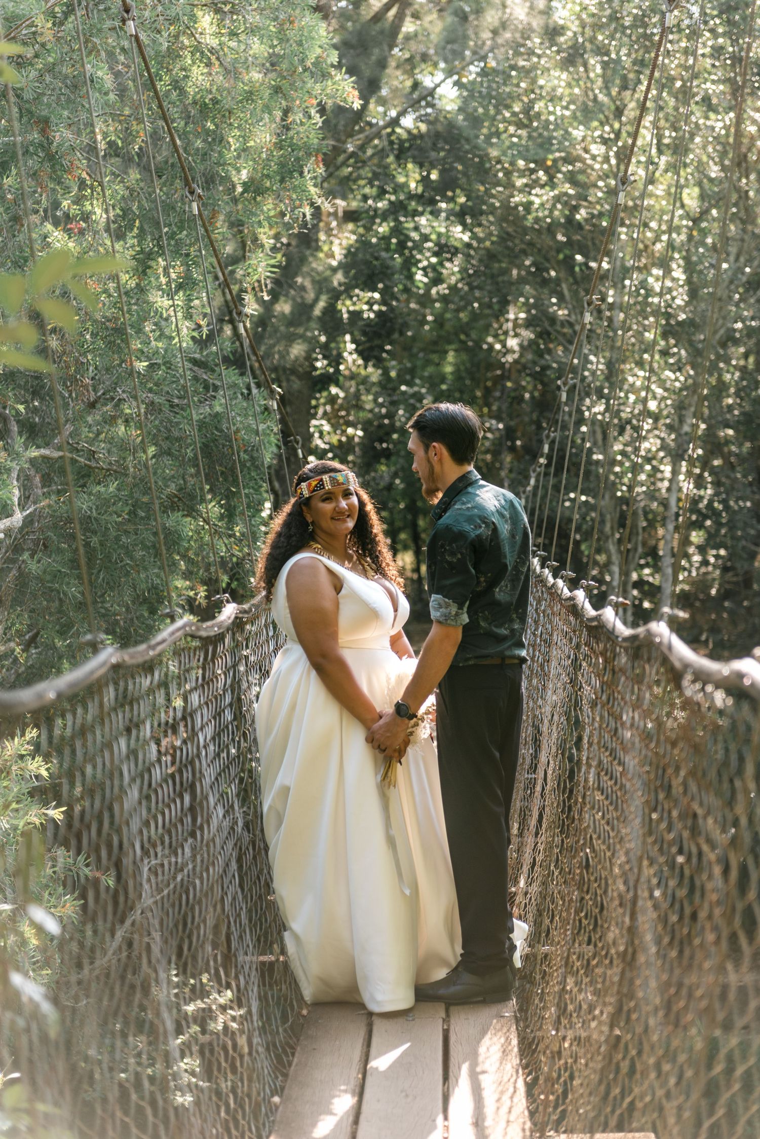 A bride and groom hold hands on a wooden suspension bridge in a lush forest setting.