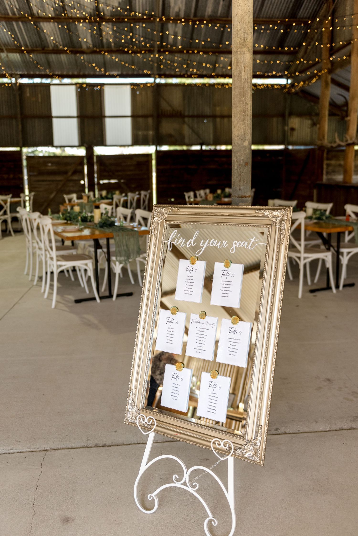 Ornate gold-framed seating chart mirror on easel in rustic barn wedding venue with fairy lights.