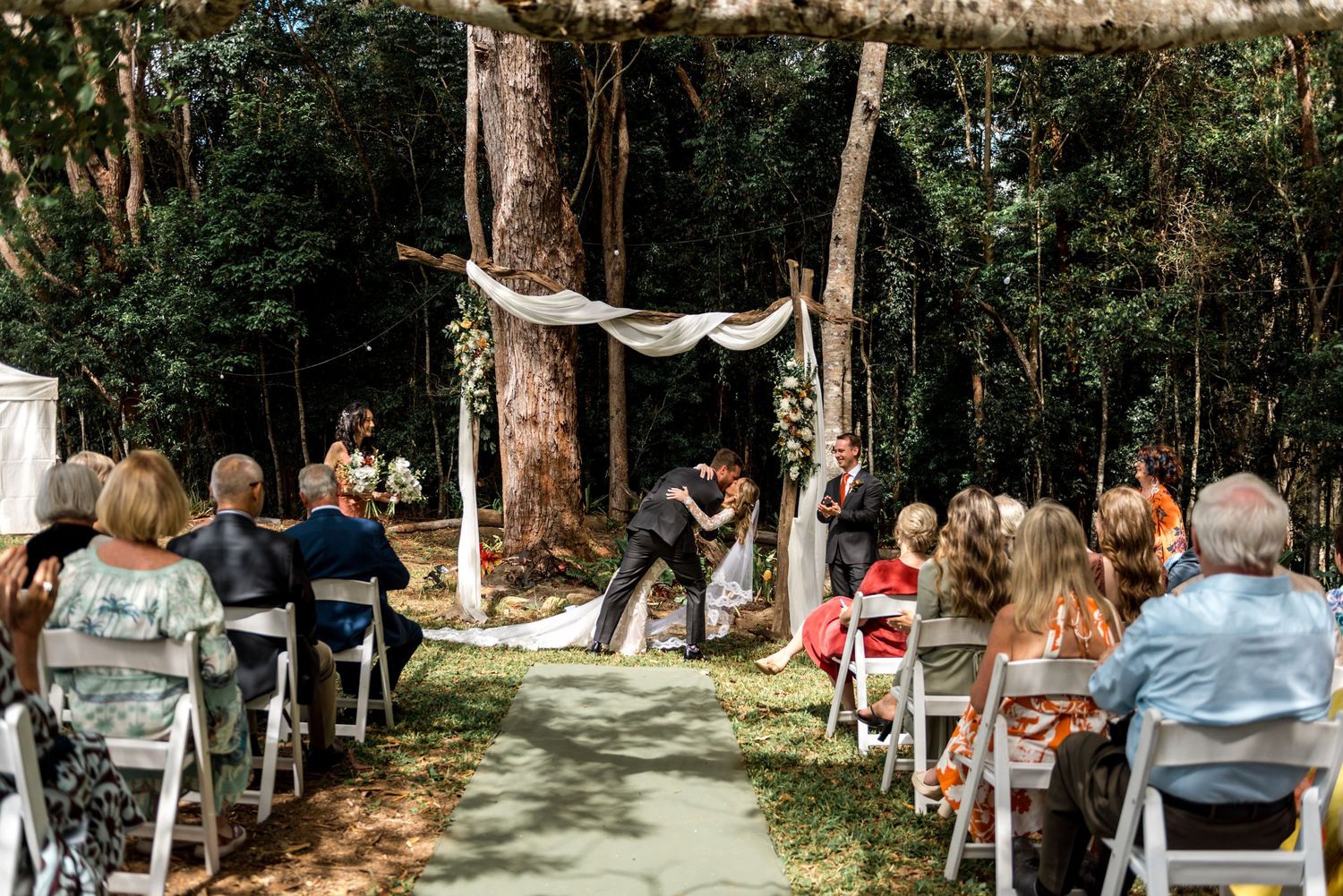 Outdoor woodland wedding ceremony with guests seated as couple exchanges vows.