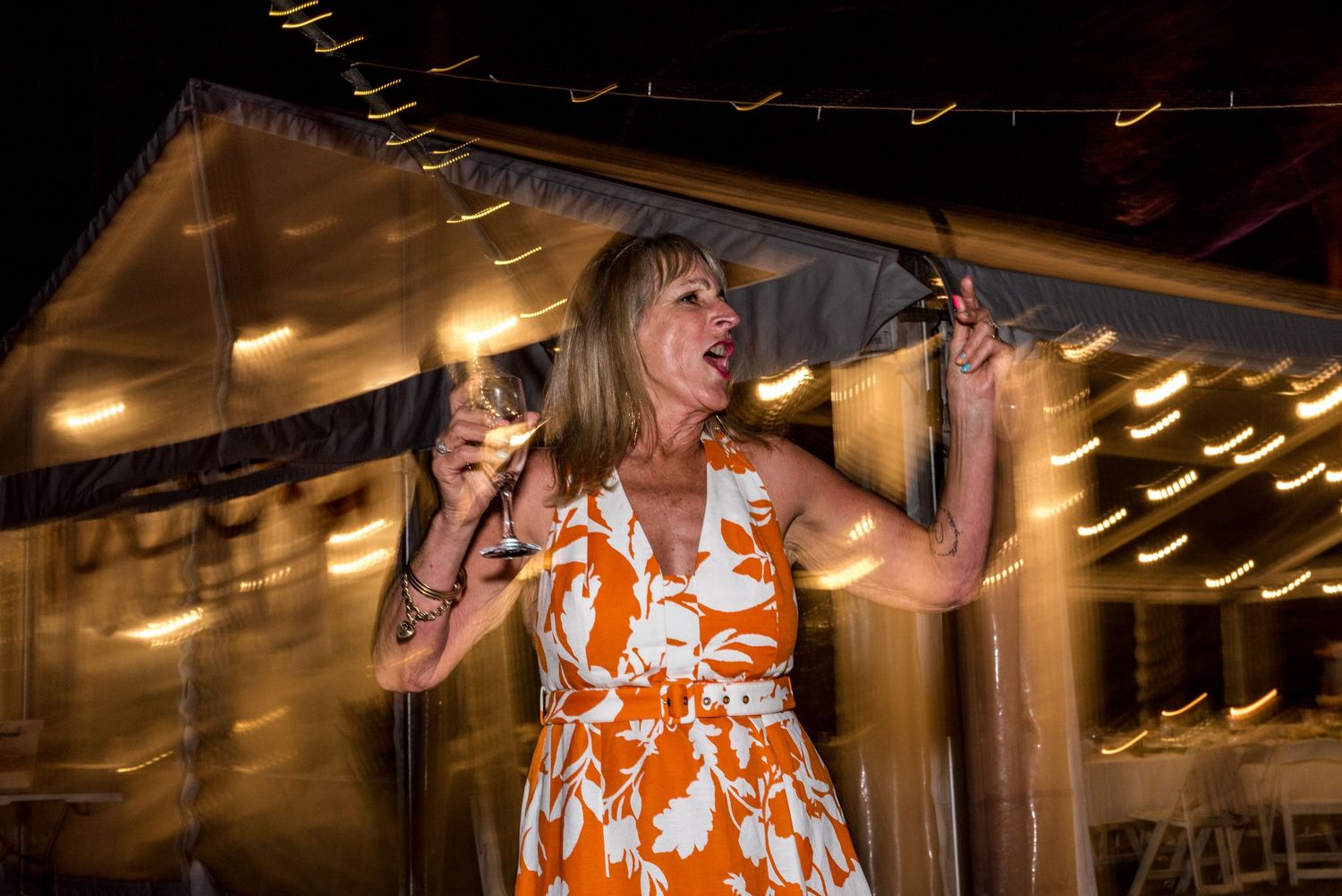Woman in orange floral dress dancing and holding wine glass at outdoor evening wedding reception.