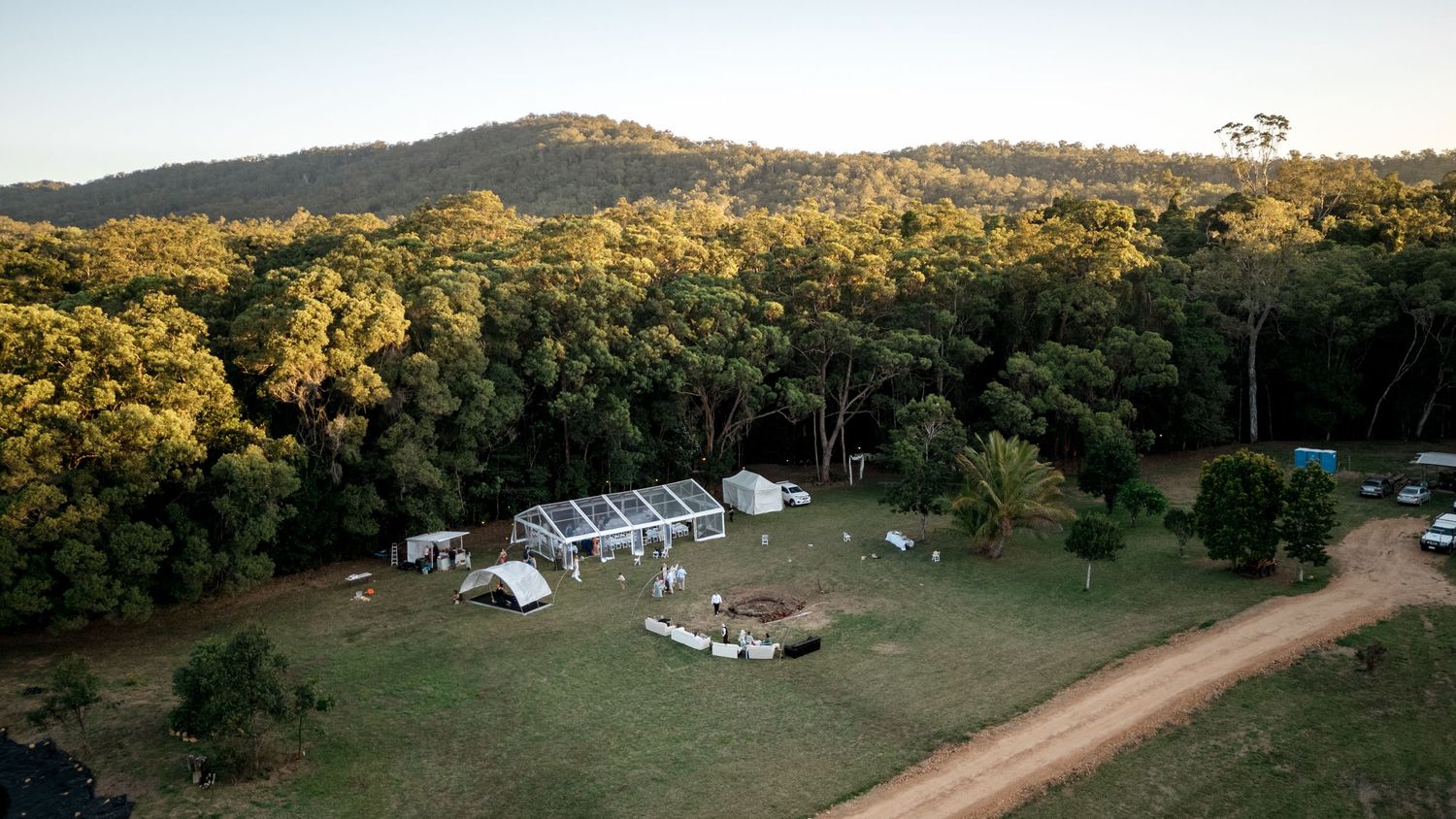 Aerial view of a rural wedding venue with marquee tent set in a forest clearing at golden hour.