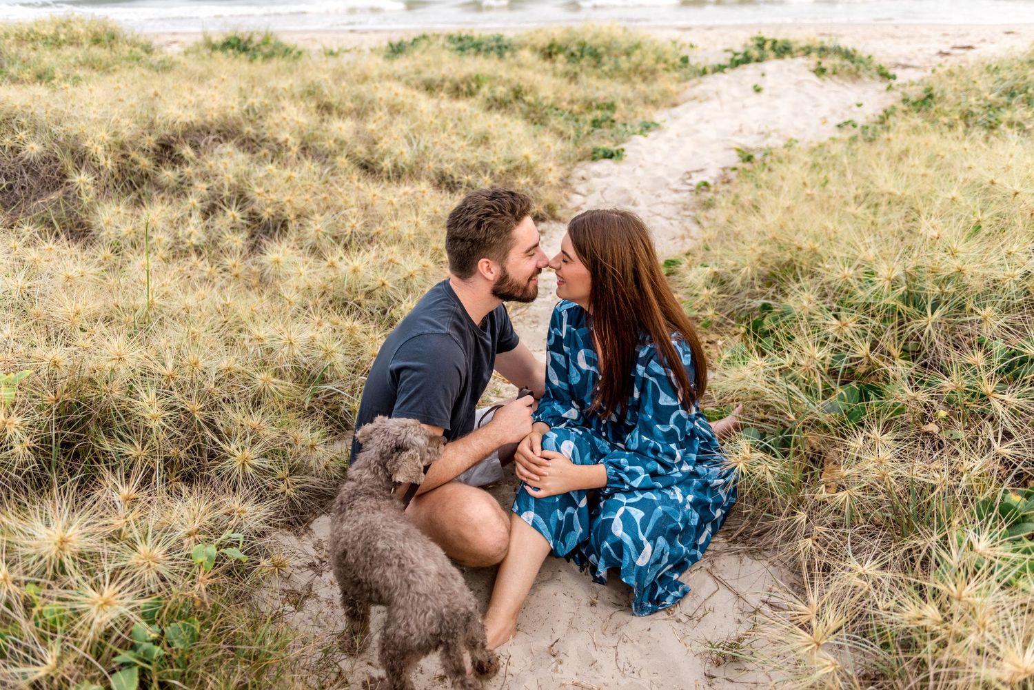 Romantic couple sitting on sand dunes about to kiss with fluffy gray dog beside them.