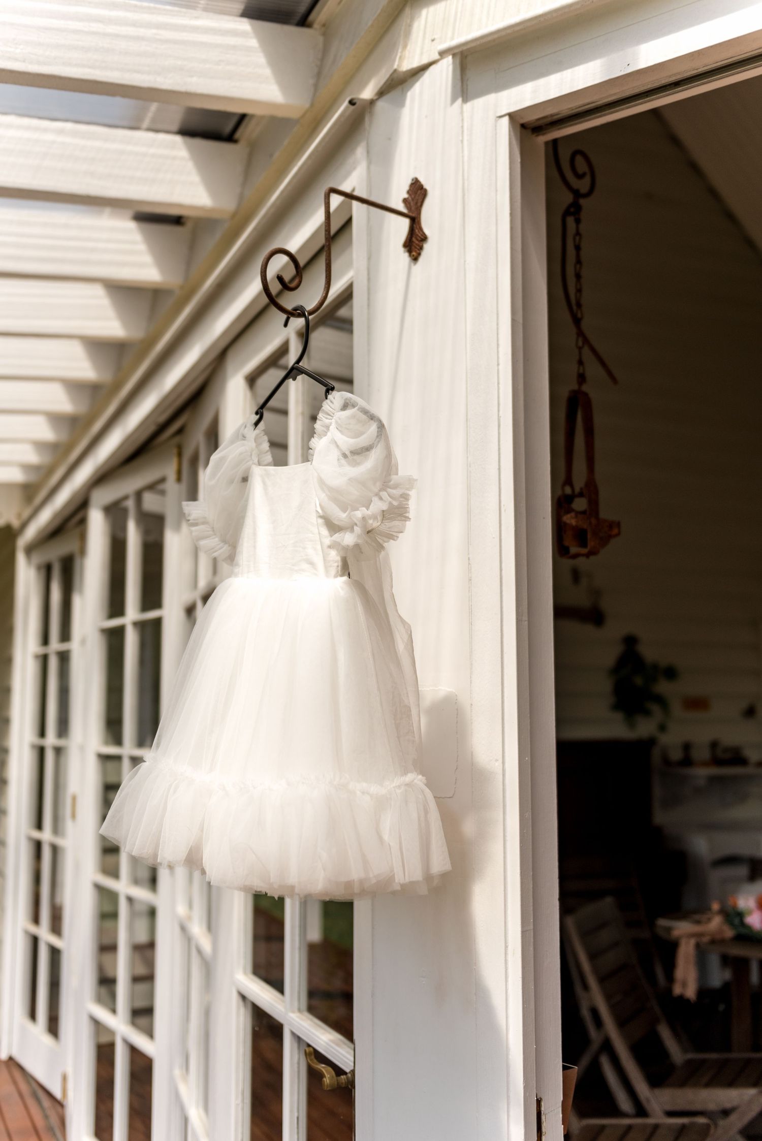 White ruffled flower girl dress hanging on ornate bracket outside a charming white timber home.