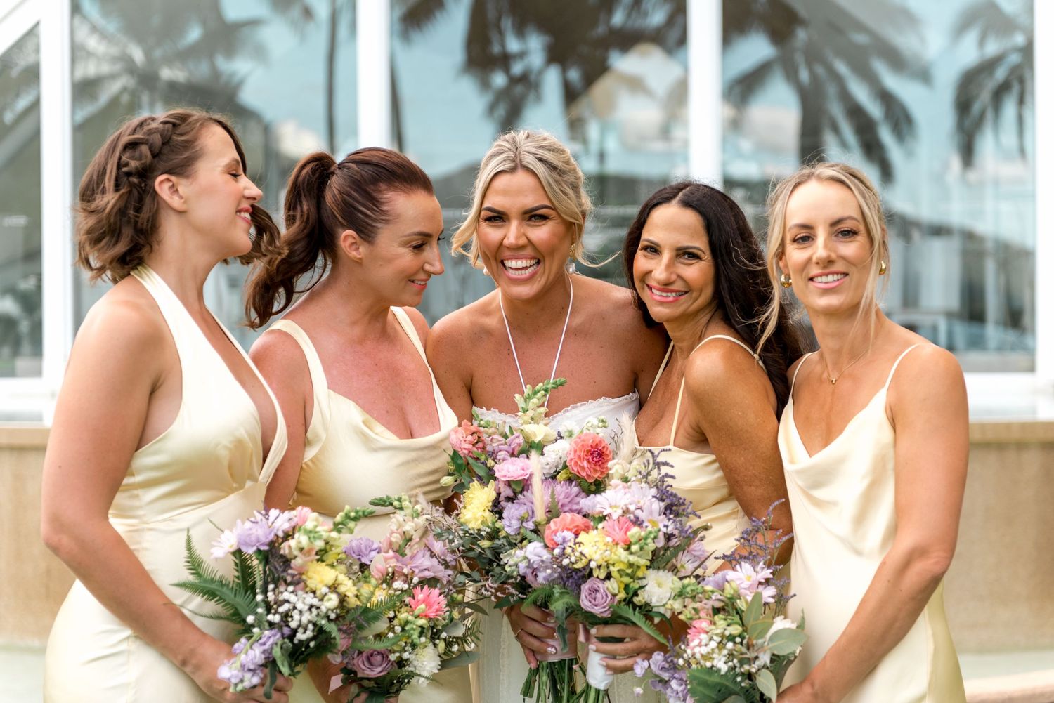 Five bridesmaids in cream dresses smiling and holding colorful bouquets at a tropical wedding.
